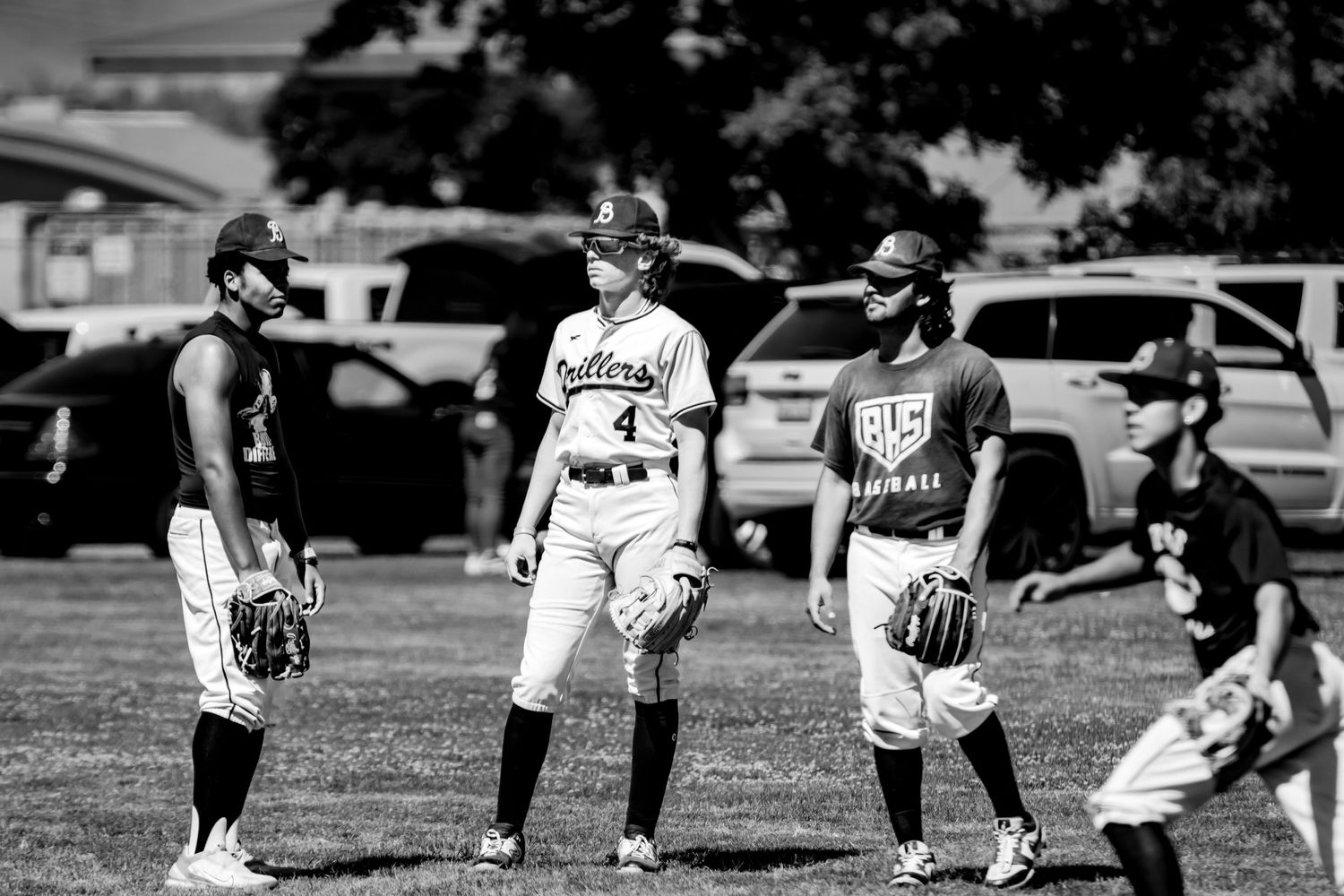 Youth baseball players interact on field during game with cars and buildings visible in distance.