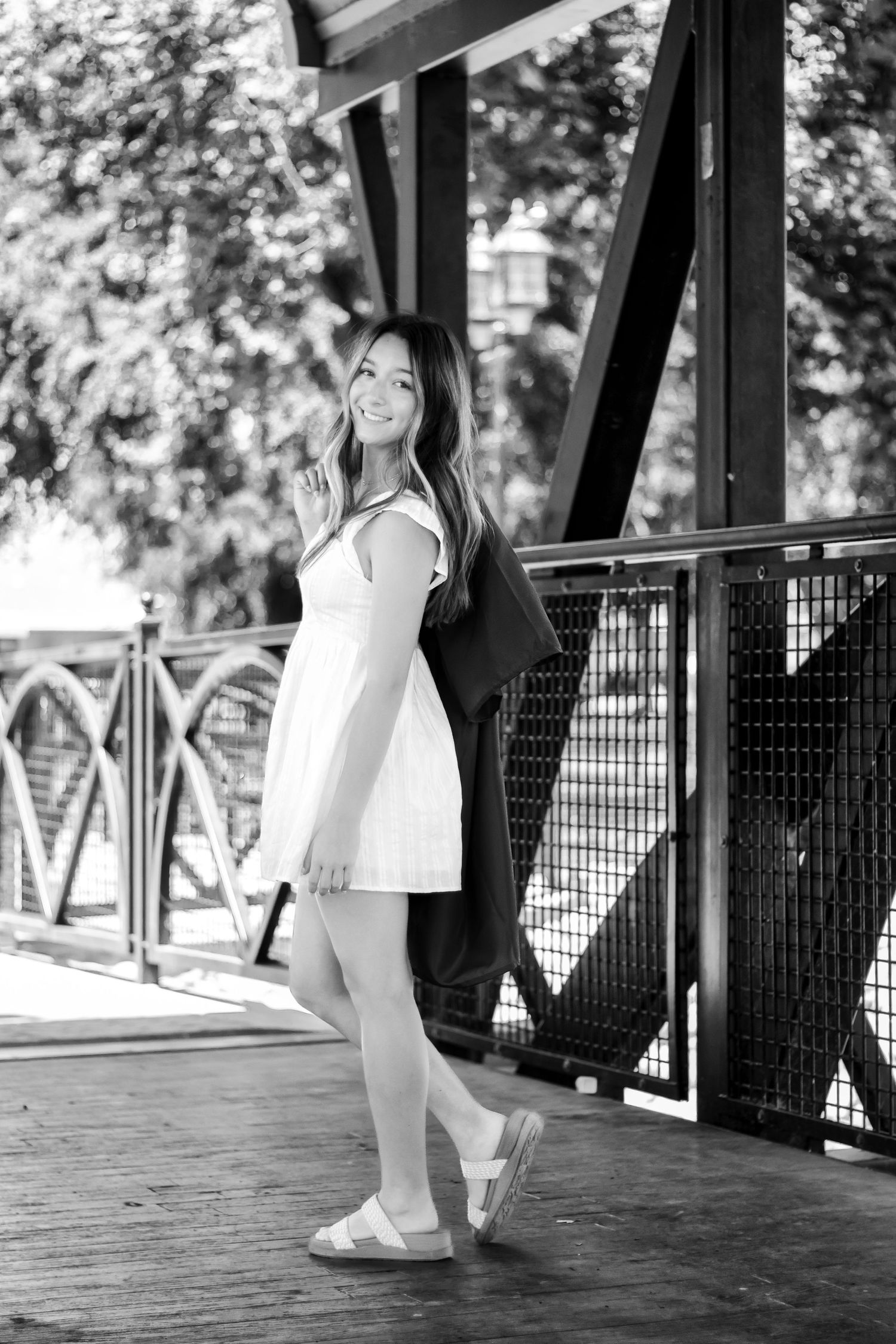 Black and white portrait of someone in a white summer dress leaning against a wooden bridge railing on a sunny day.
