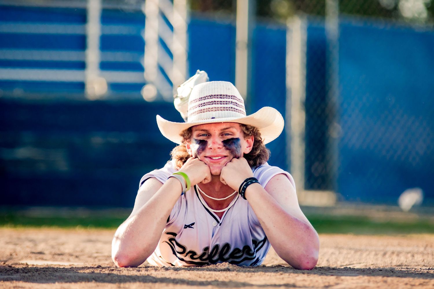 A baseball player in a white uniform and cowboy hat lies on the ground making a silly face at the camera.