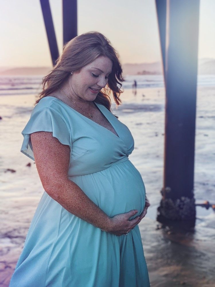 A pregnant person in a light blue dress stands beneath a pier at sunset on the beach.