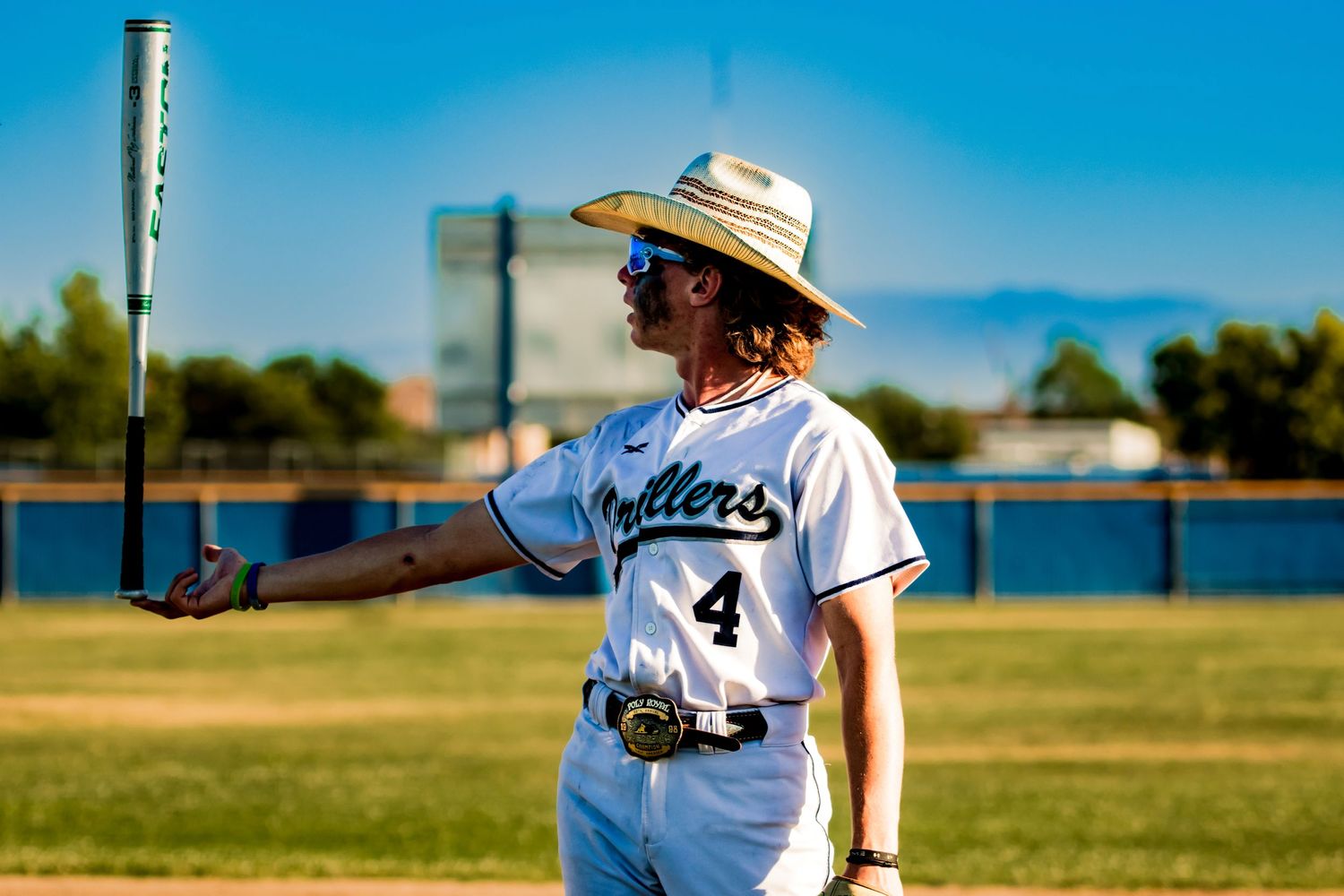 Baseball player in white uniform and straw hat holding bat on sunny field.