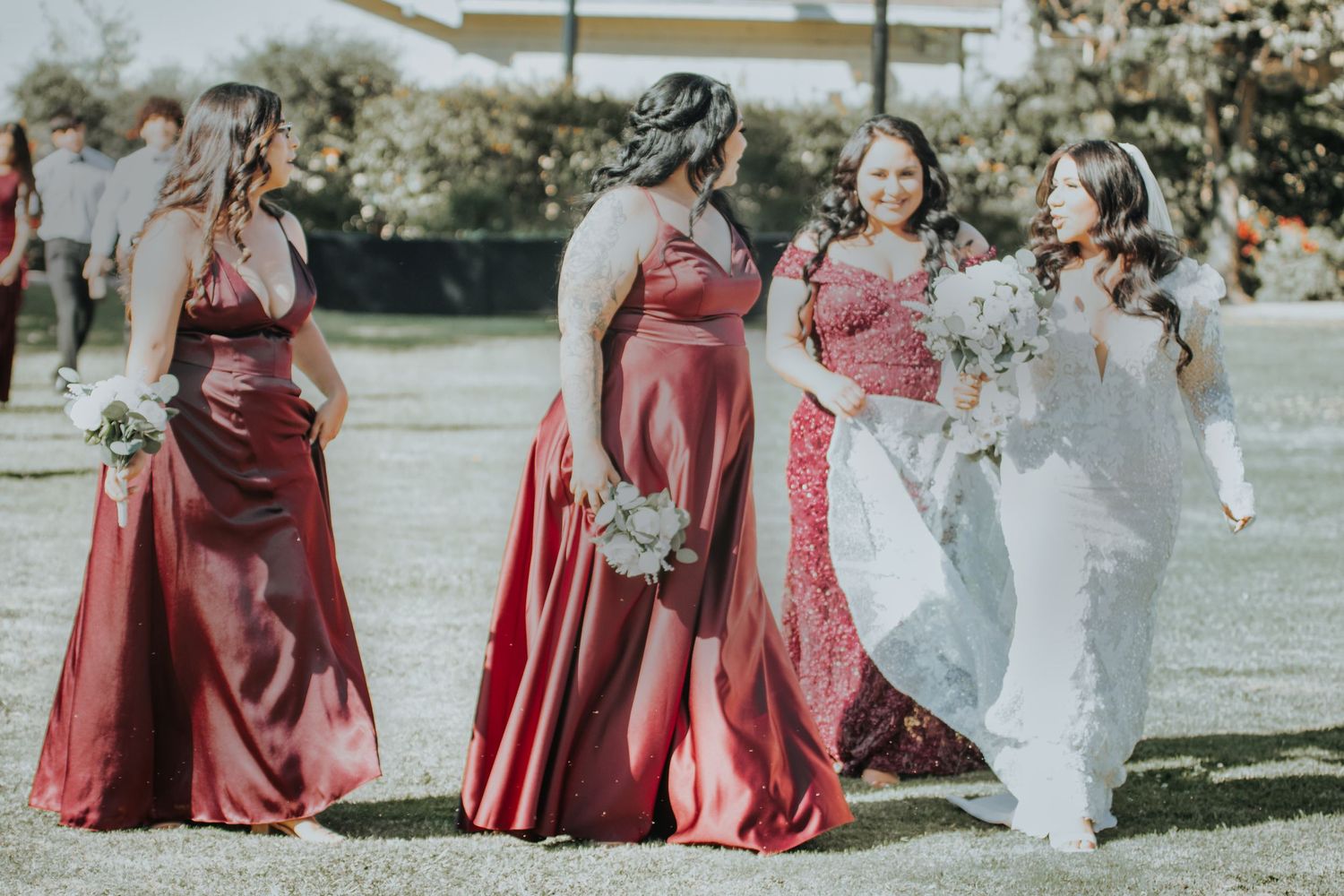 Bridesmaids in flowing red gowns stand with bride in white dress on sunny lawn during outdoor wedding celebration.