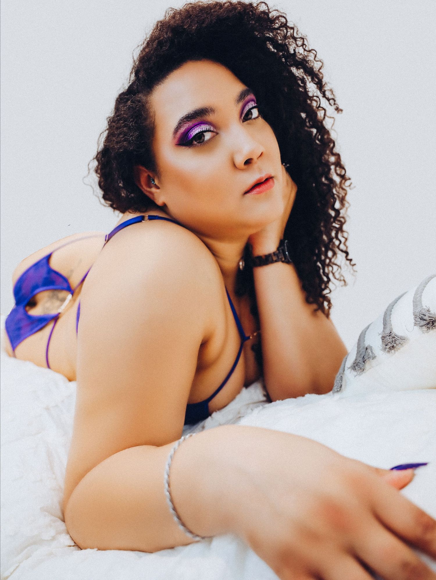 Boudoir portrait of confident and beautiful woman in purple lingerie on white bedding for photoshoot with Boudoir by Bri Saint Louis