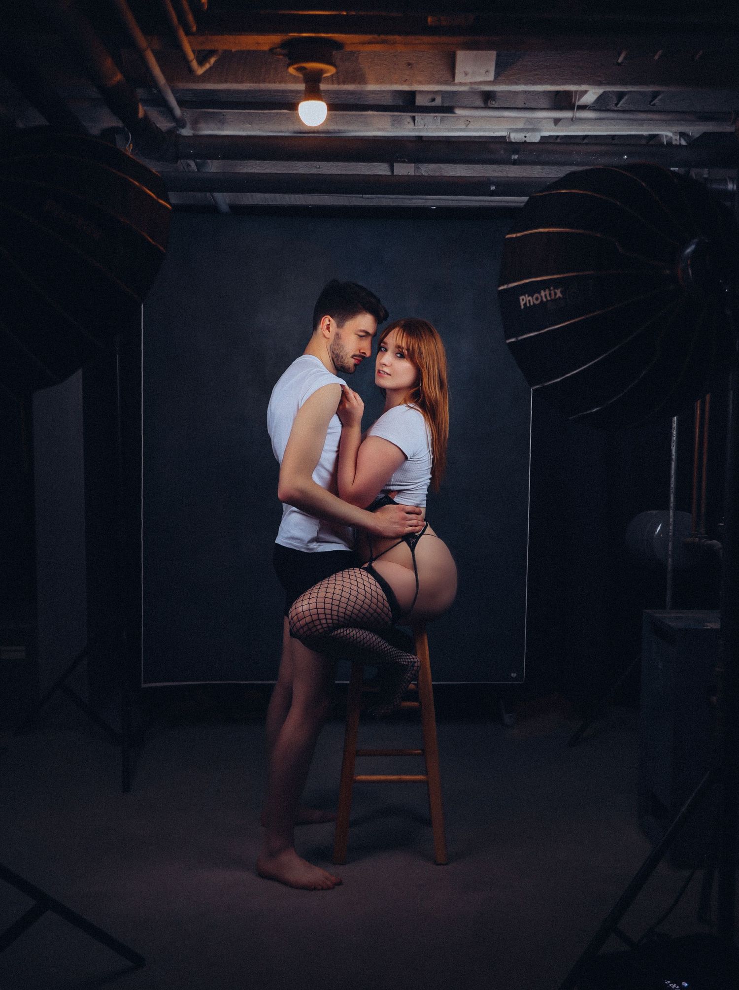 Boudoir portrait of flirty young couple in love holding each other close in front of blue backdrop for photoshoot with Boudoir by Bri