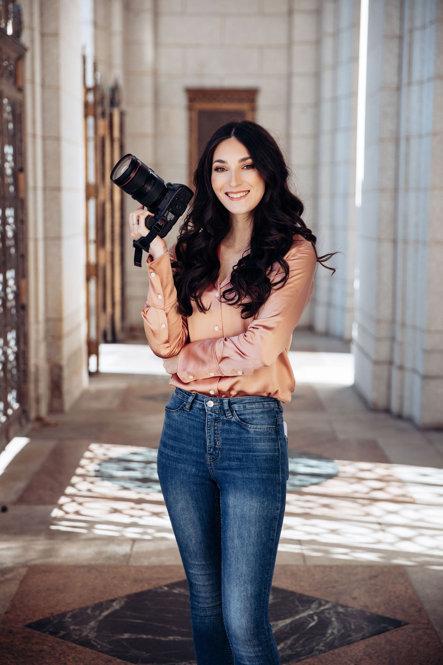 Photographer portrait of woman with long black curled hair wearing pink silk shirt smiling and holding professional camera in Saint Louis