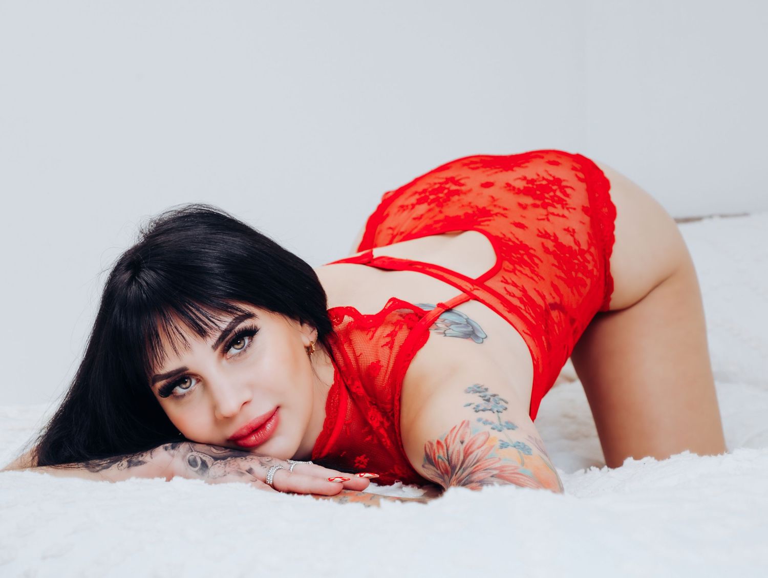 Beautiful woman with tattoos and long black hair and bangs posing in white bedroom wearing red lingerie piece for Boudoir by Bri Saint Louis