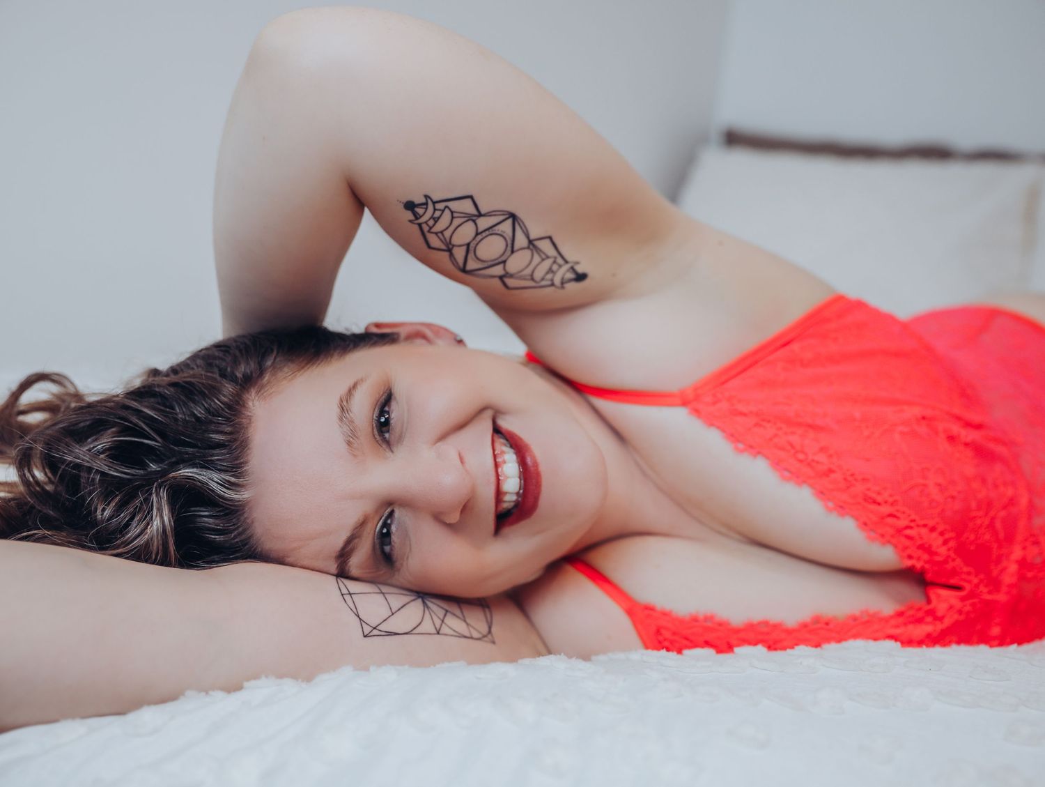 Woman laying on side, smiling into camera wearing hot pink lingerie on white bed for photoshoot with Boudoir by Bri in Saint Louis