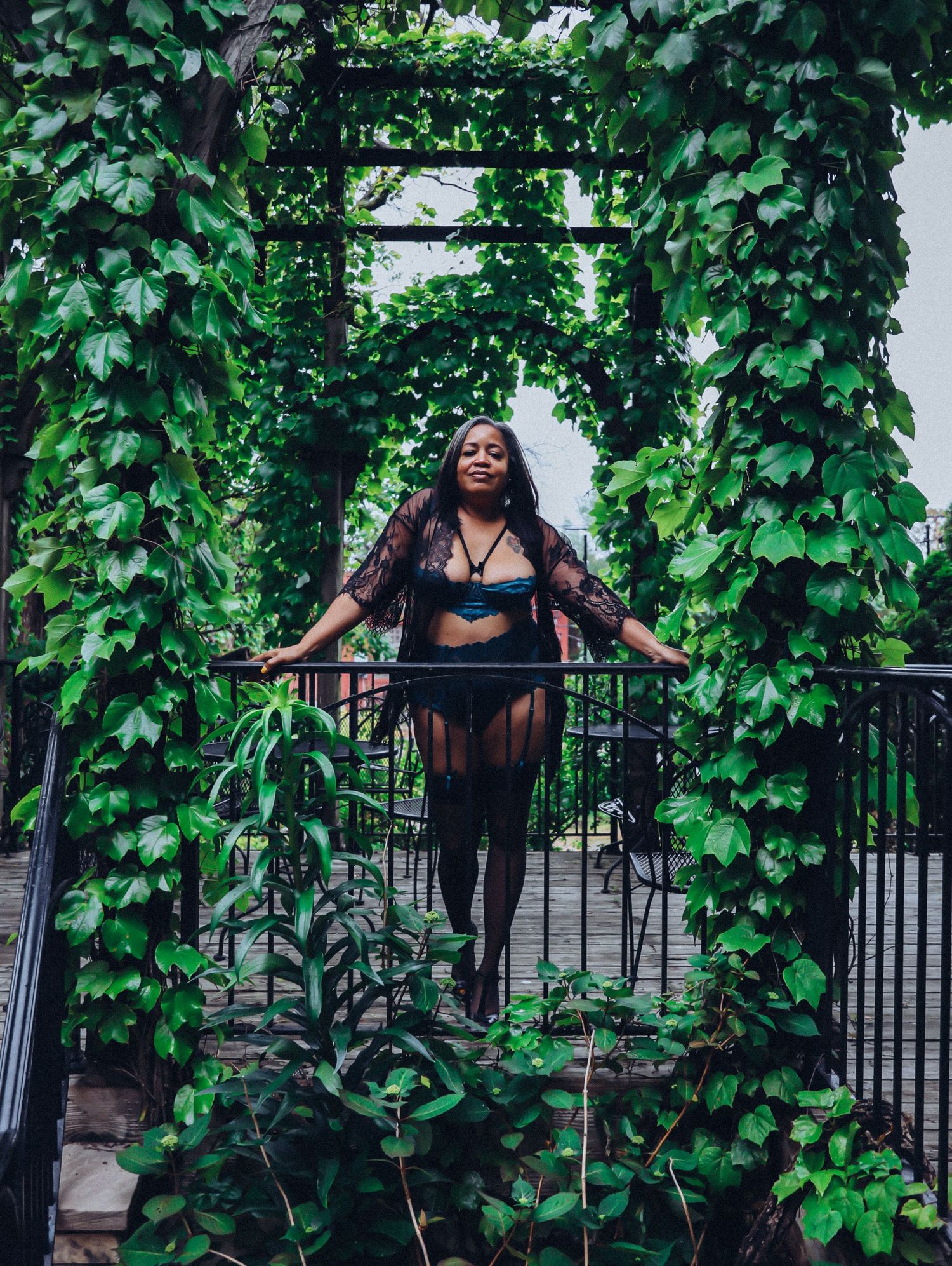 Beautiful woman in black lingerie set leaning on gate surrounded by thick greenery for photoshoot Boudoir by Bri in Saint Louis
