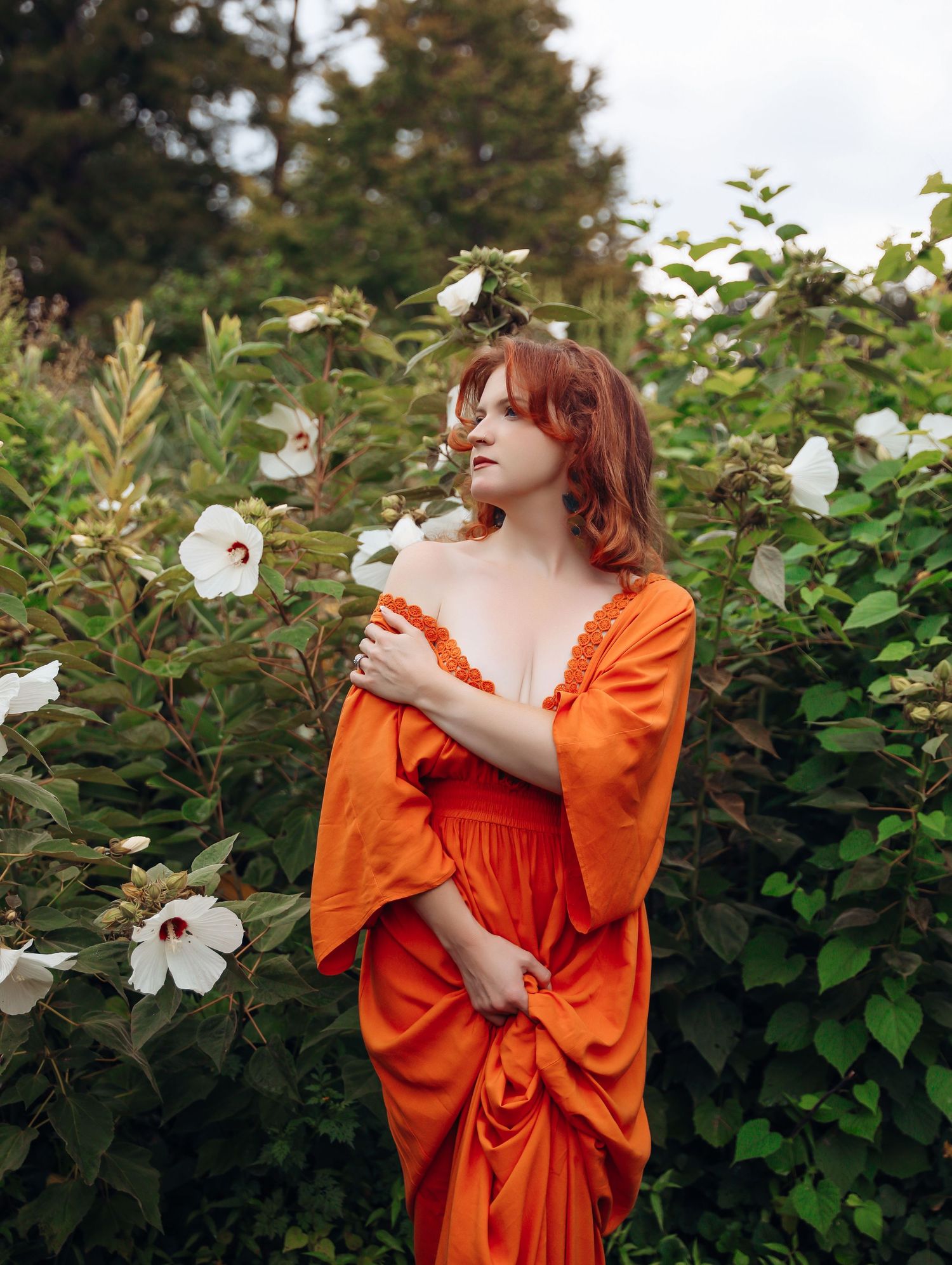 Woman with curly red hair wearing beautiful orange flowy gown posing in greenery for outdoor photoshoot with Boudoir by Bri St. Louis
