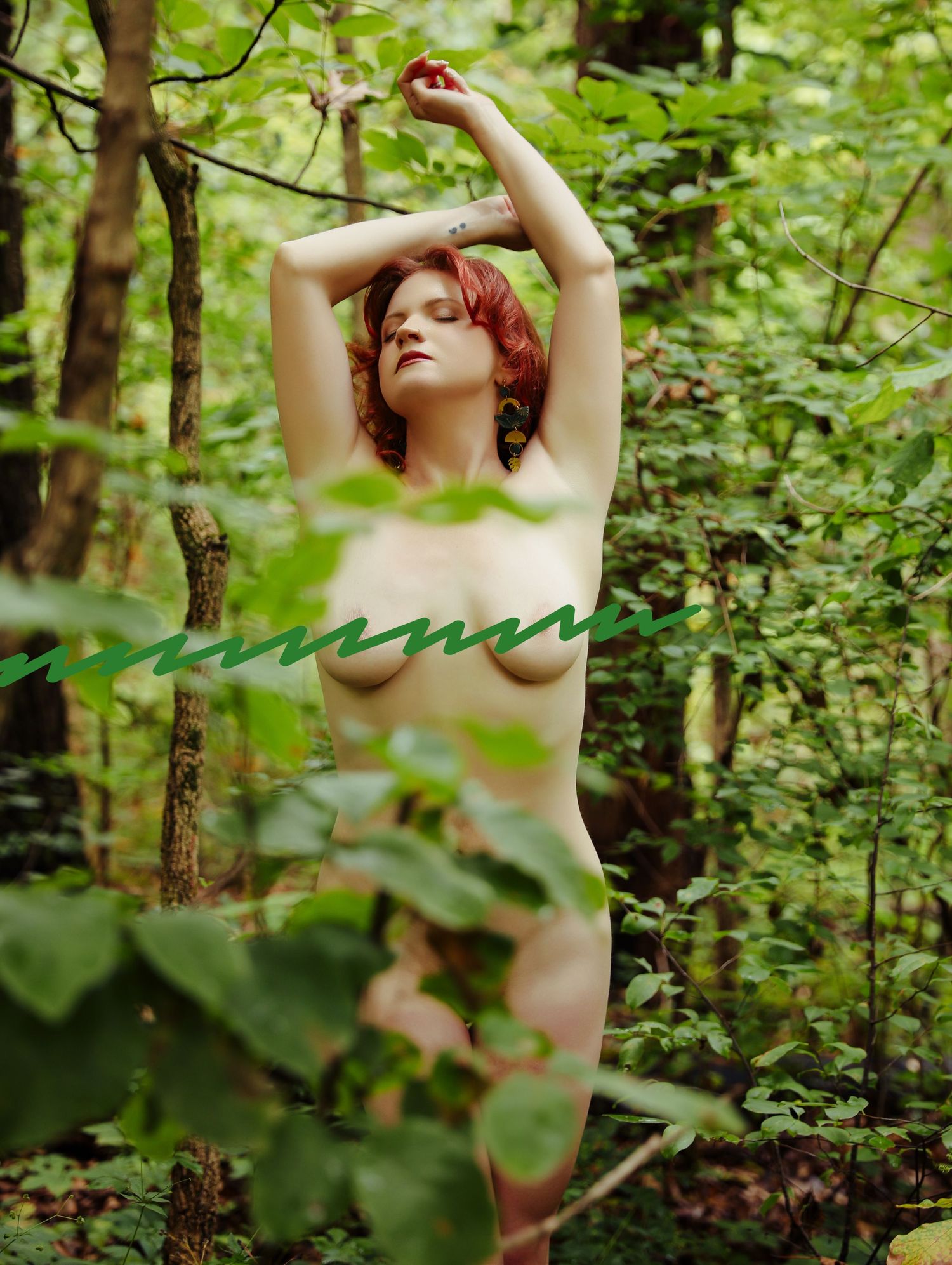 Woman with curly red hair posing completely naked in woods surrounded by greenery for outdoor shoot with Boudoir by Bri in Saint Louis