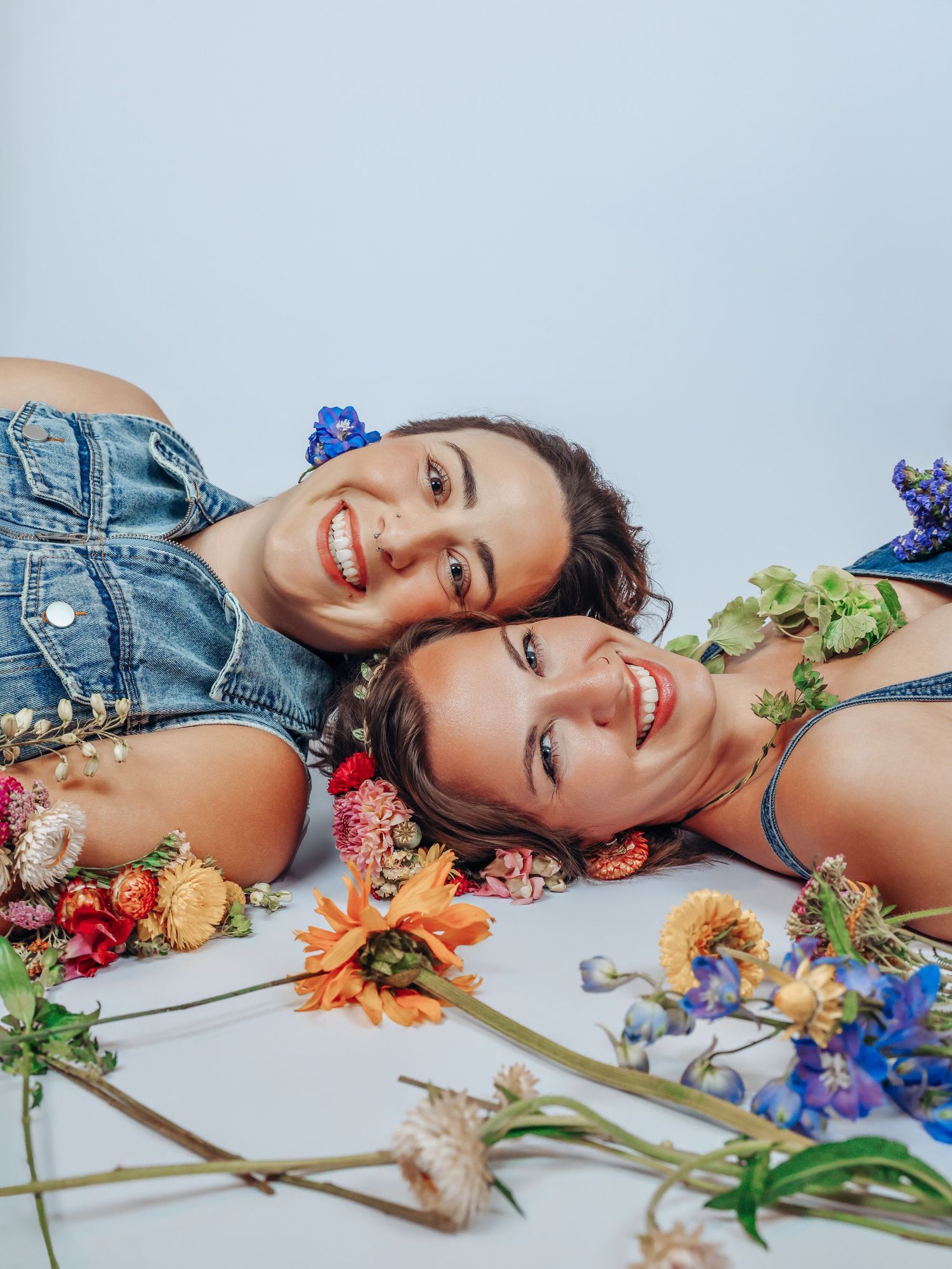 Best friends on white backdrop, heads touching, wild flowers scattered for photoshoot with Boudoir by Bri in St. Louis Missouri