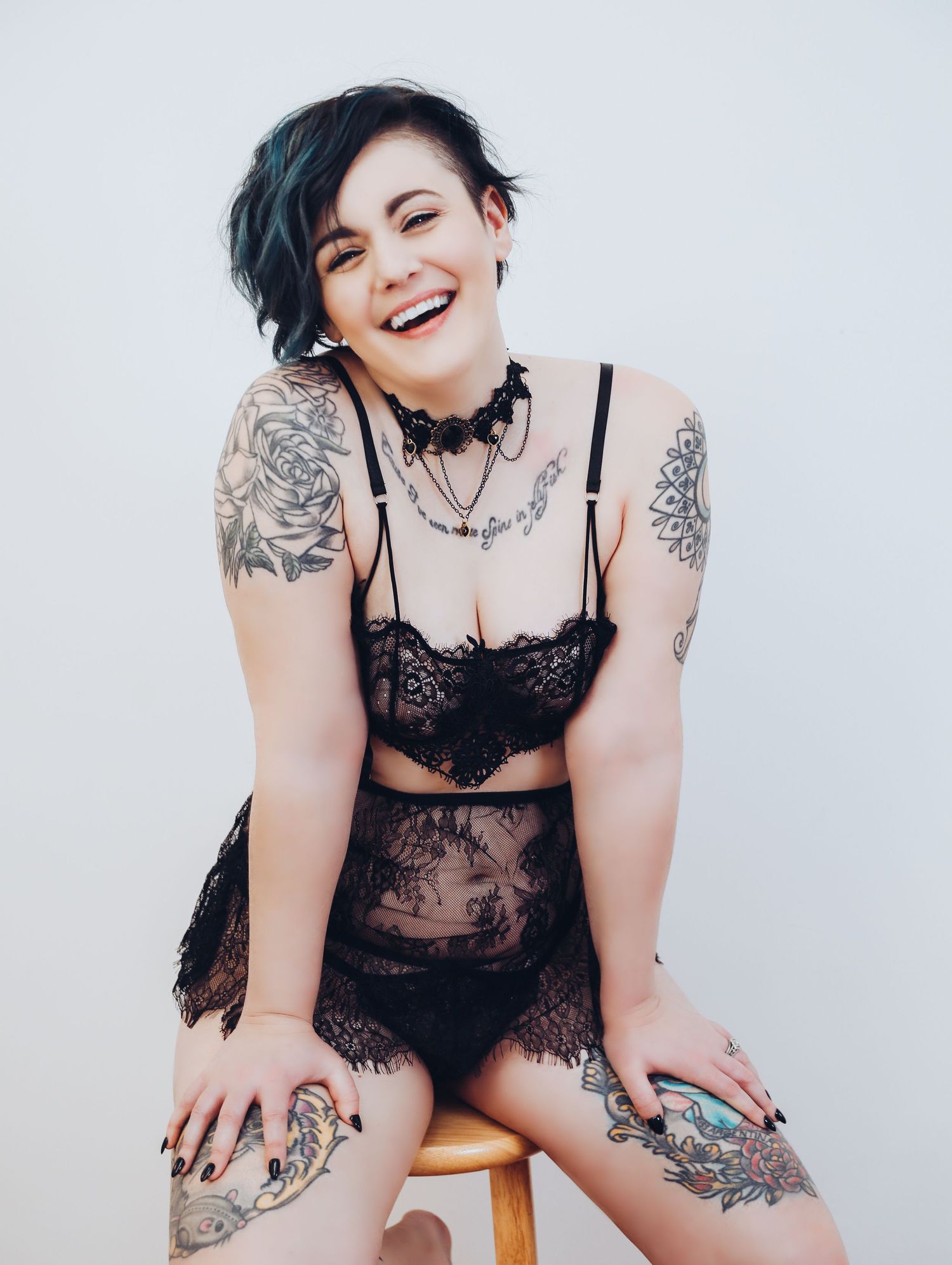 Beautiful woman with tattoos and short black and blue hair laughing into camera on wooden stool for photoshoot with Boudoir by Bri STL