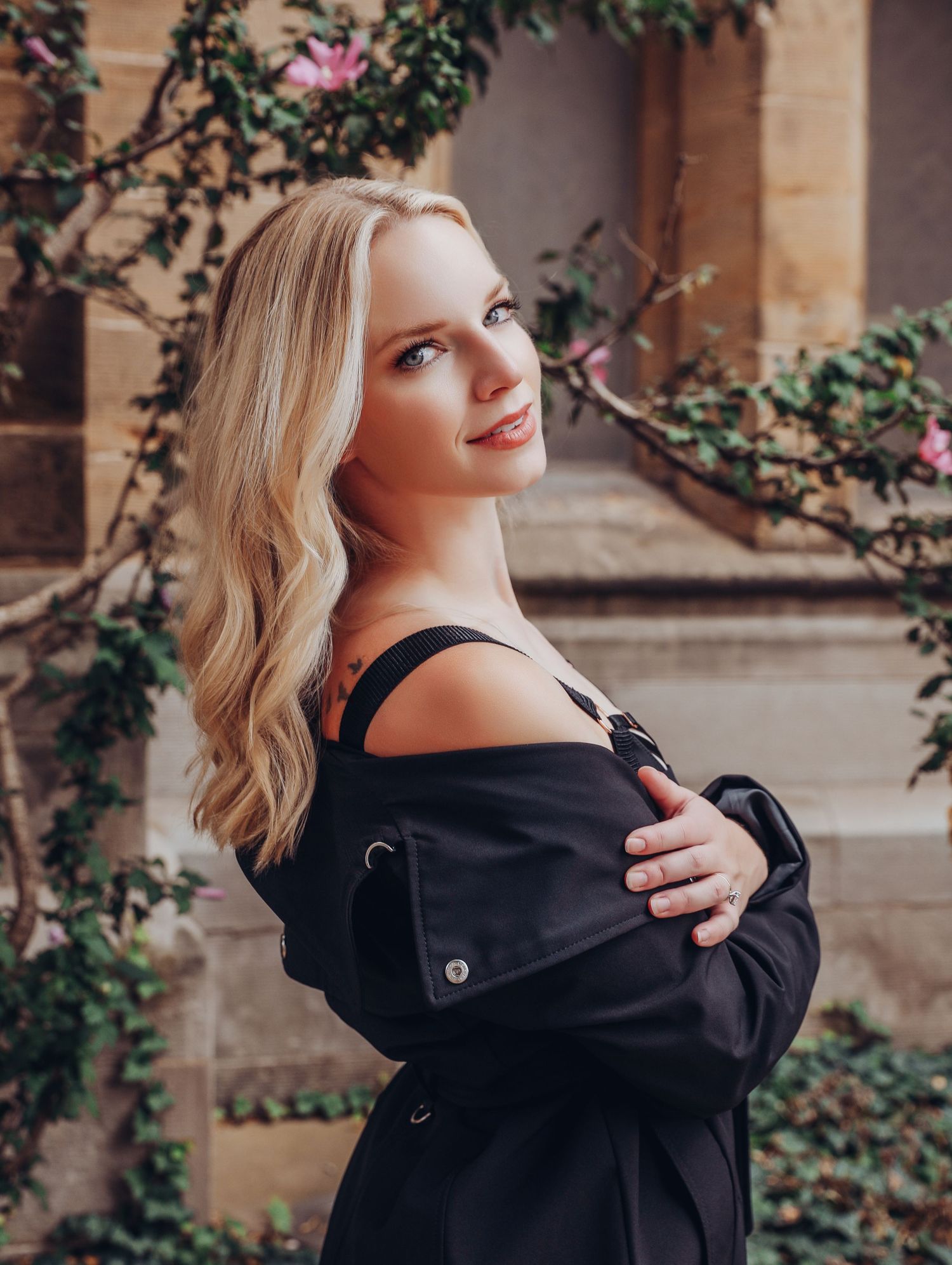 Gorgeous blonde woman posing in front of pink blossoms in black trench coat with lingerie underneath for Boudoir by Bri in Saint Louis