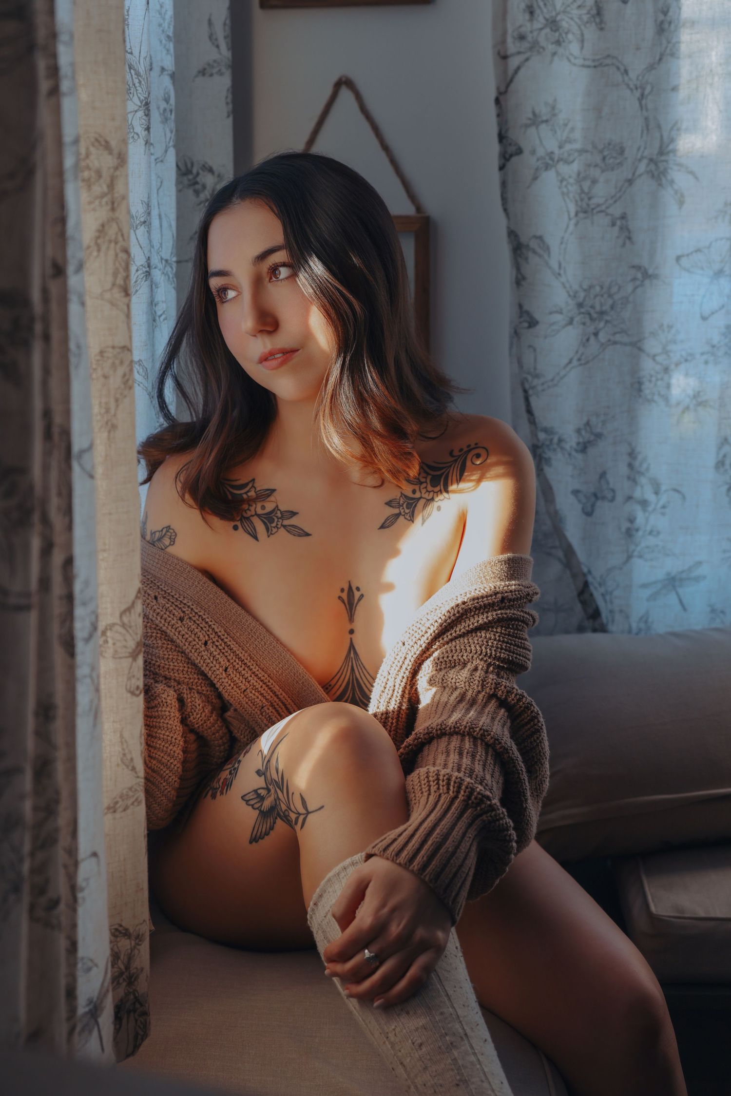 Beautiful woman with tattoos and sweater posing by window evening sunset natural light for photoshoot with Boudoir by Bri in Saint Louis MO
