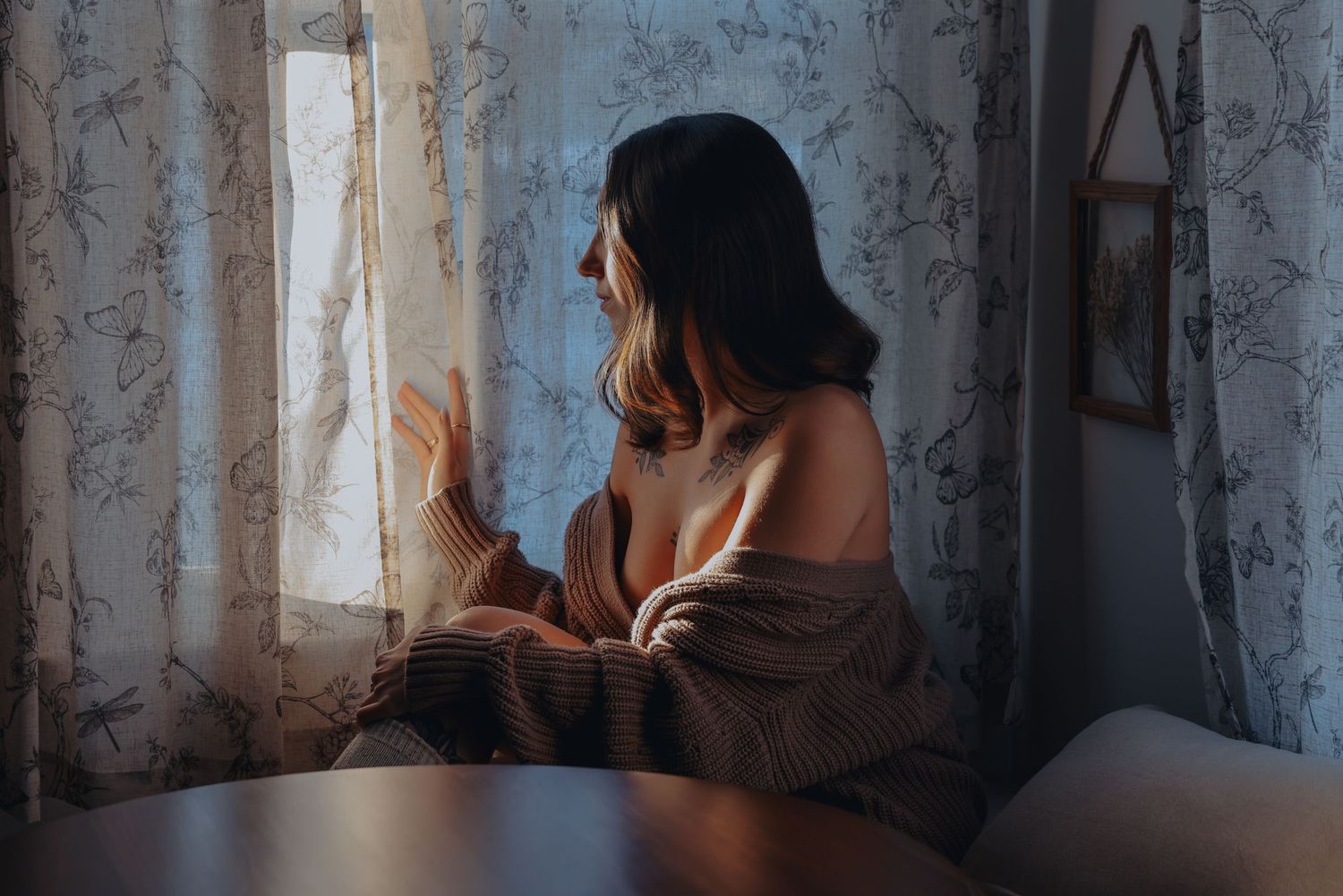 Beautiful young woman at kitchen table looking through curtain, brown sweater falling off shoulders for photoshoot, Boudoir by Bri St. Louis