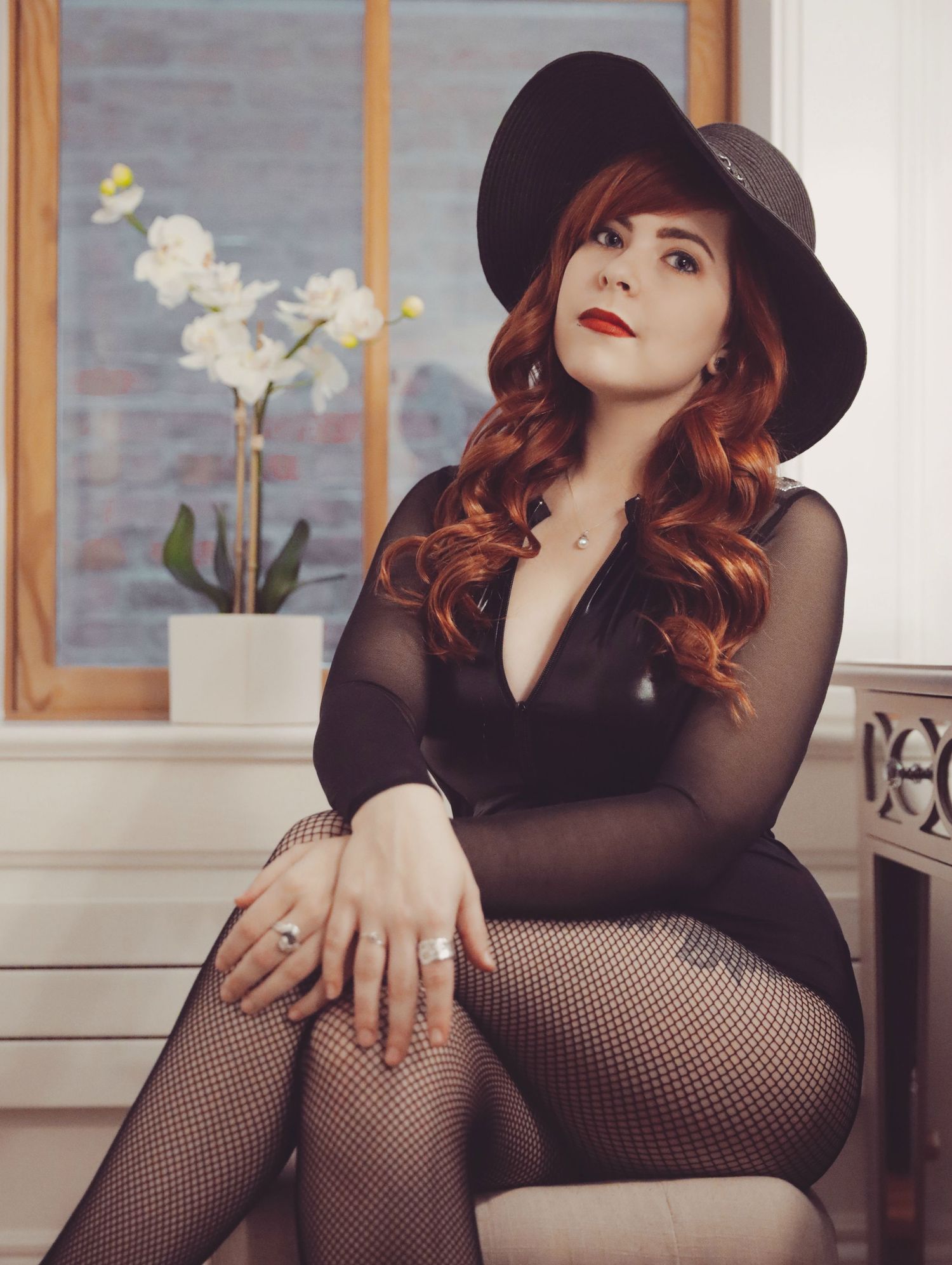 Woman with long, red, curled hair posing on ottoman in front of window with orchid in background, wearing black fishnets, leotard, and hat.