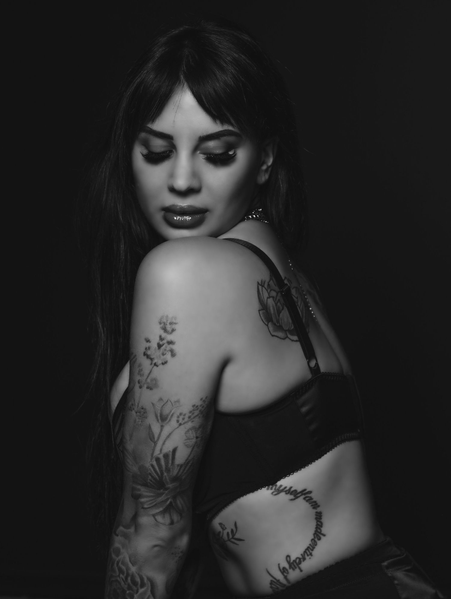Beautiful black and white portrait of young woman in bra with tattoos and black hair with bangs, looking over shoulder eyes closed St. Louis