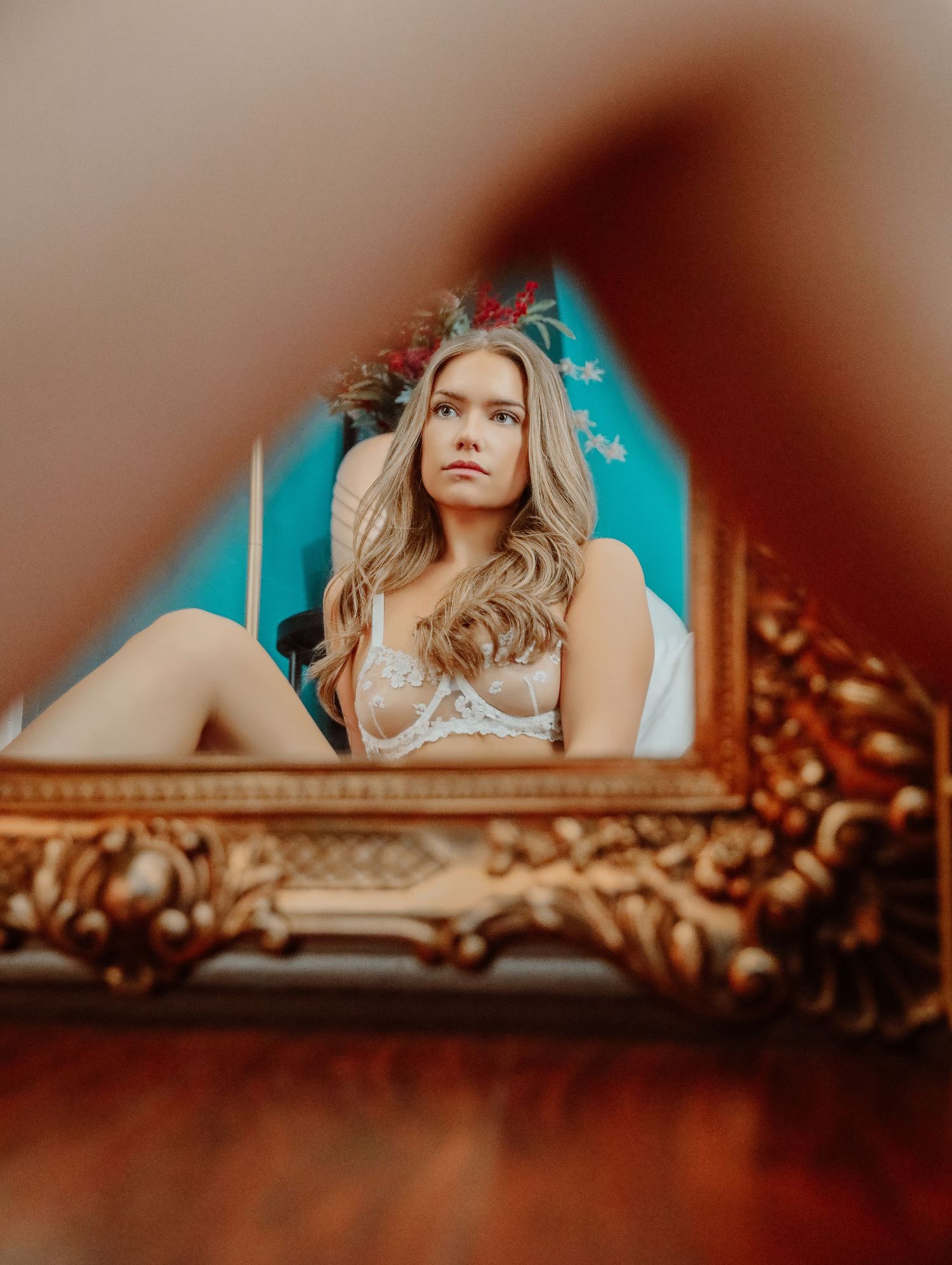 Blonde woman in white bridal boudoir lingerie sitting on floor looking at herself in reflection of gold, guided mirror