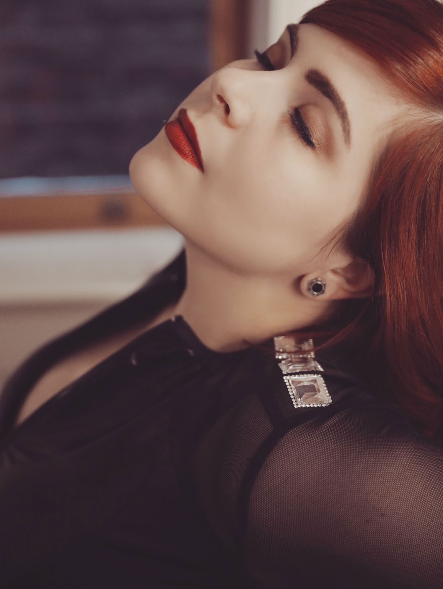 Beautiful portrait of pale woman with red hair and red lipstick wearing black, eyes closed and face turned up to ceiling.