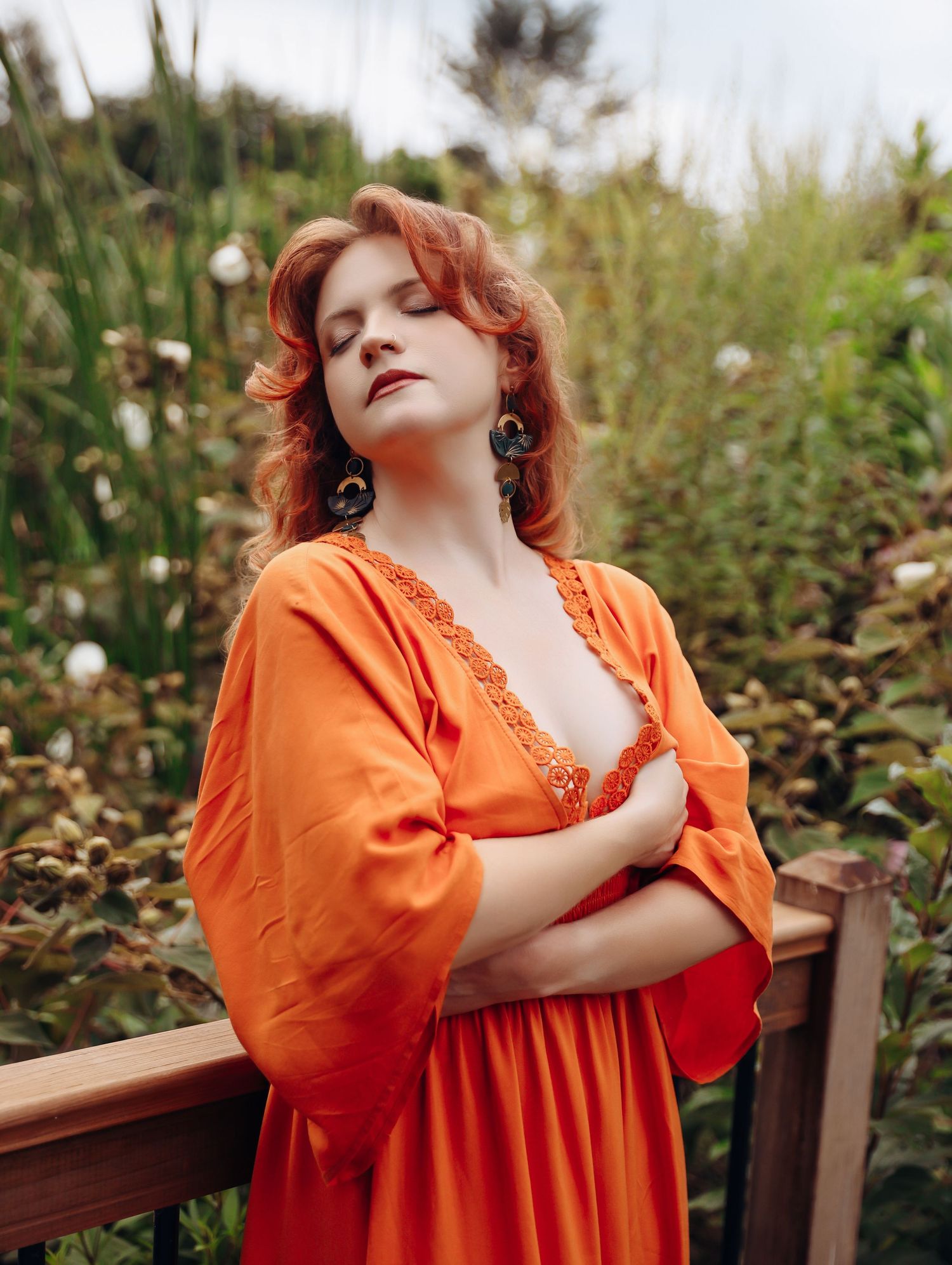 Woman with curly red hair wearing beautiful orange flowy gown posing in greenery for outdoor photoshoot with Boudoir by Bri St. Louis