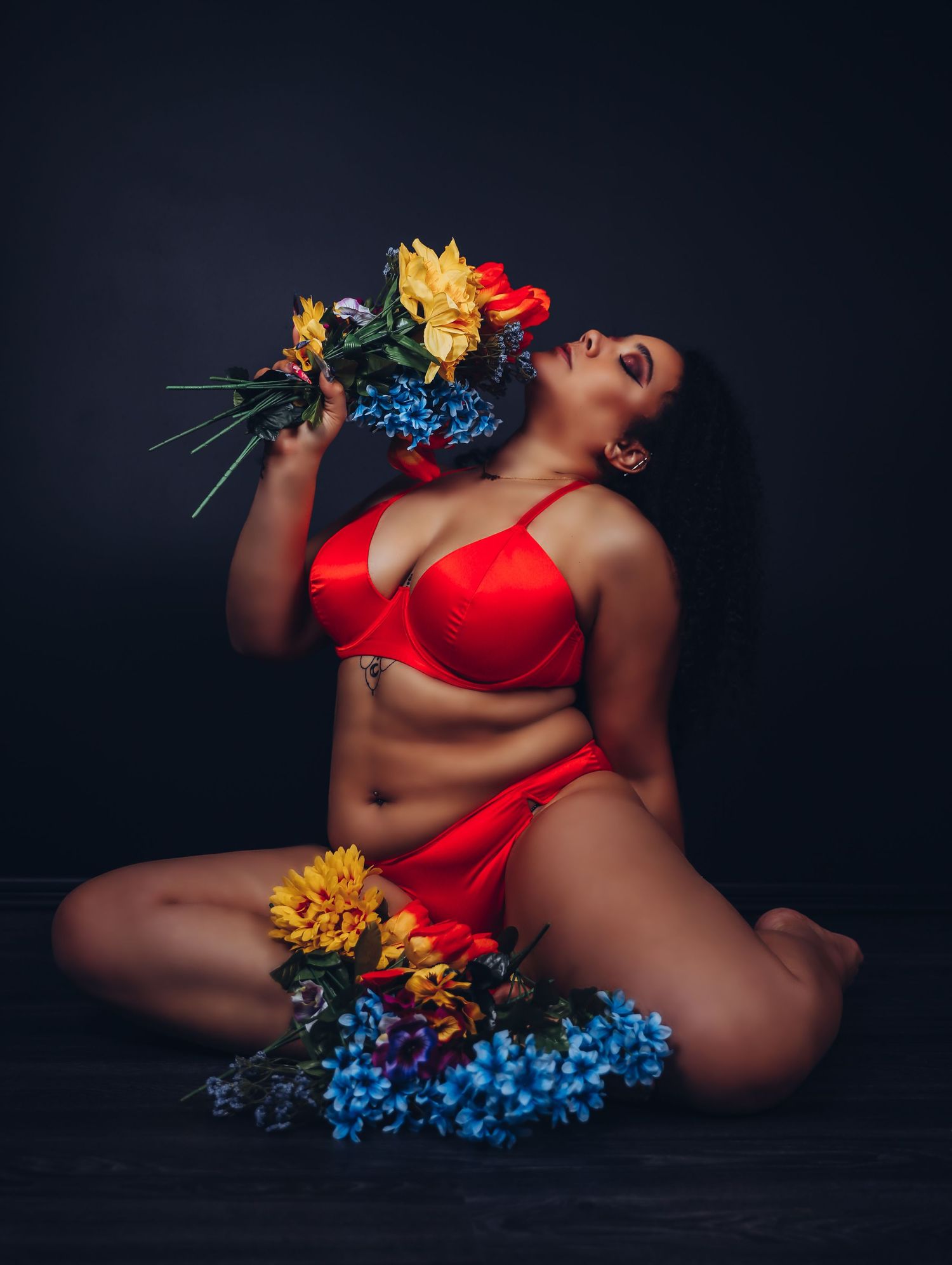 Dramatic boudoir portrait of beautiful woman in orange lingerie set holding spring flowers against black background Boudoir by Bri St. Louis