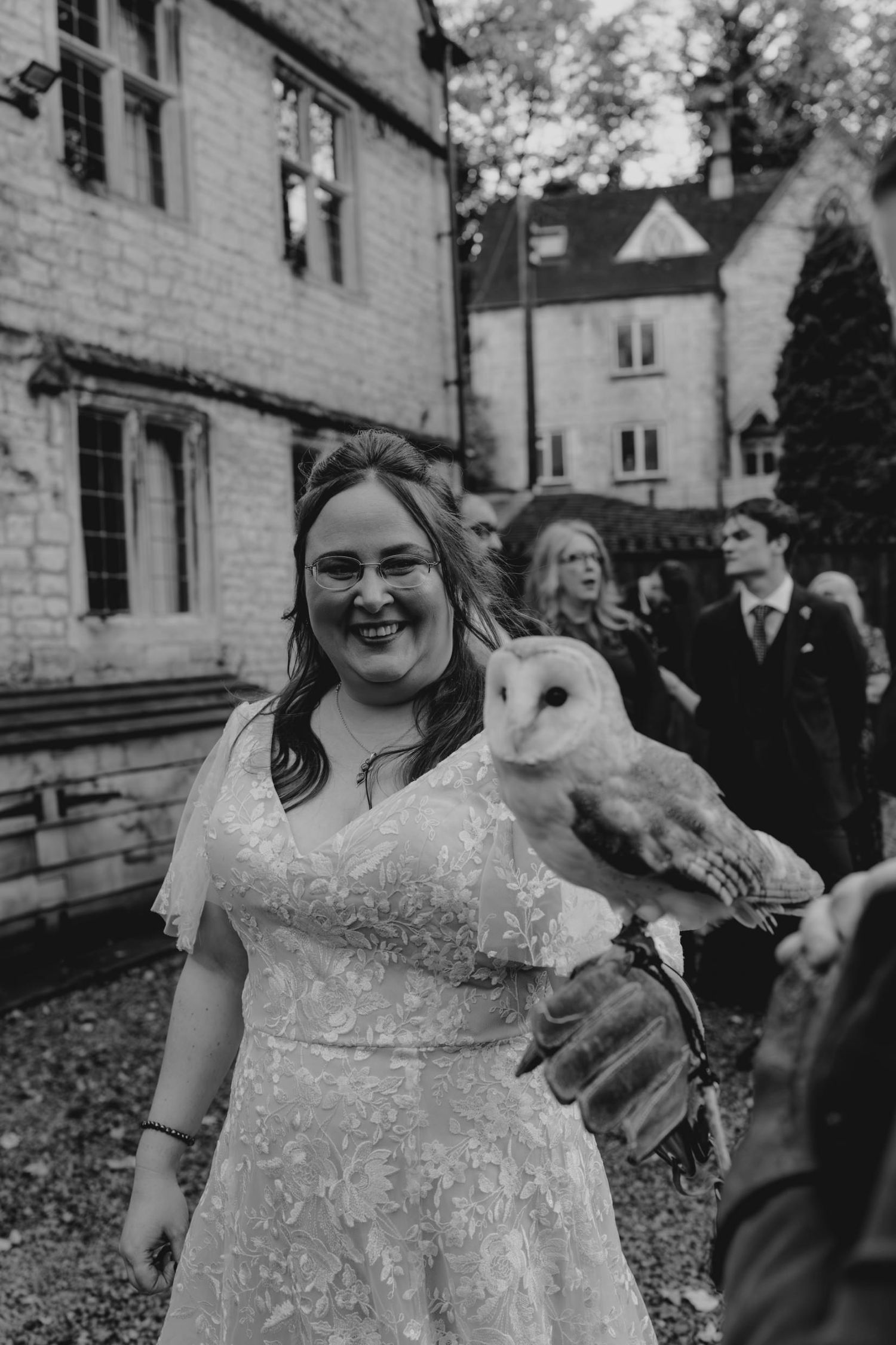 Smiling bride in lace wedding dress holds a barn owl outside a stone manor house in black and white photo.