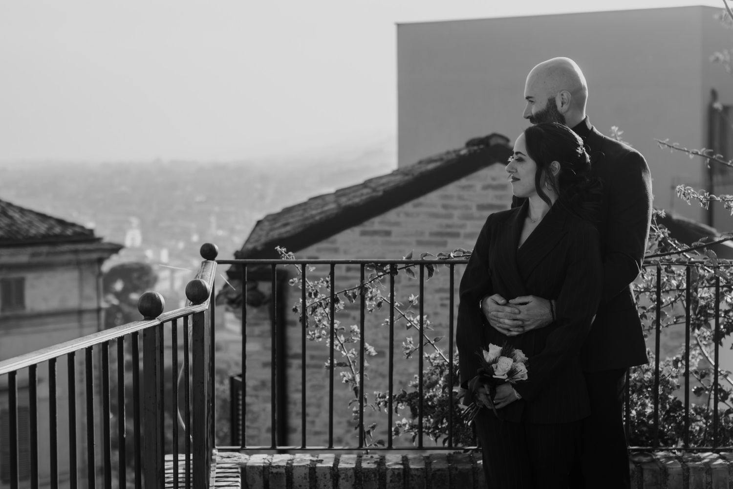 Elegant couple in black attire embracing on a balcony with a cityscape backdrop in a black and white photo.