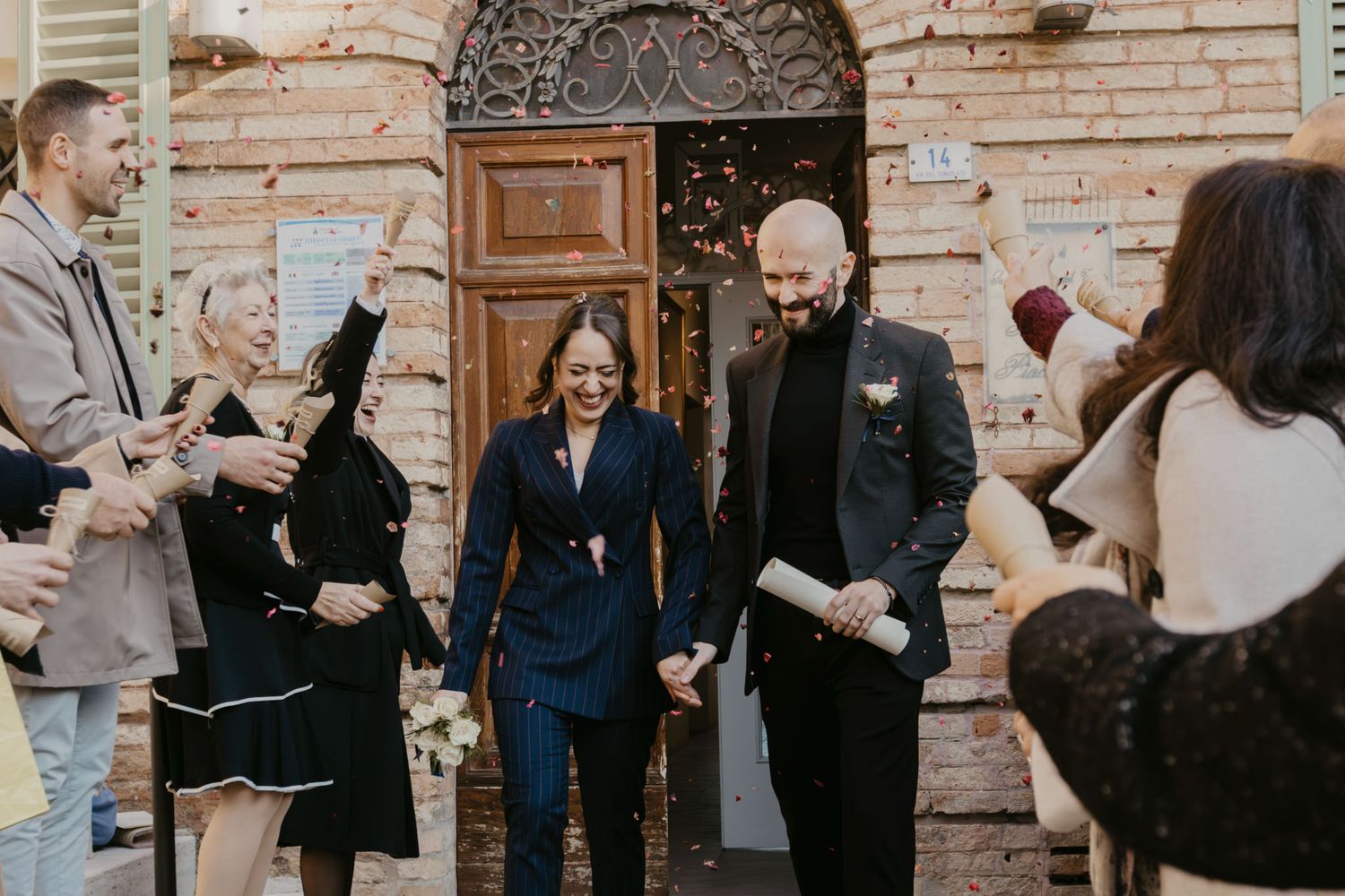 Newlywed couple exits a rustic Italian building amid confetti, surrounded by smiling guests in elegant attire.