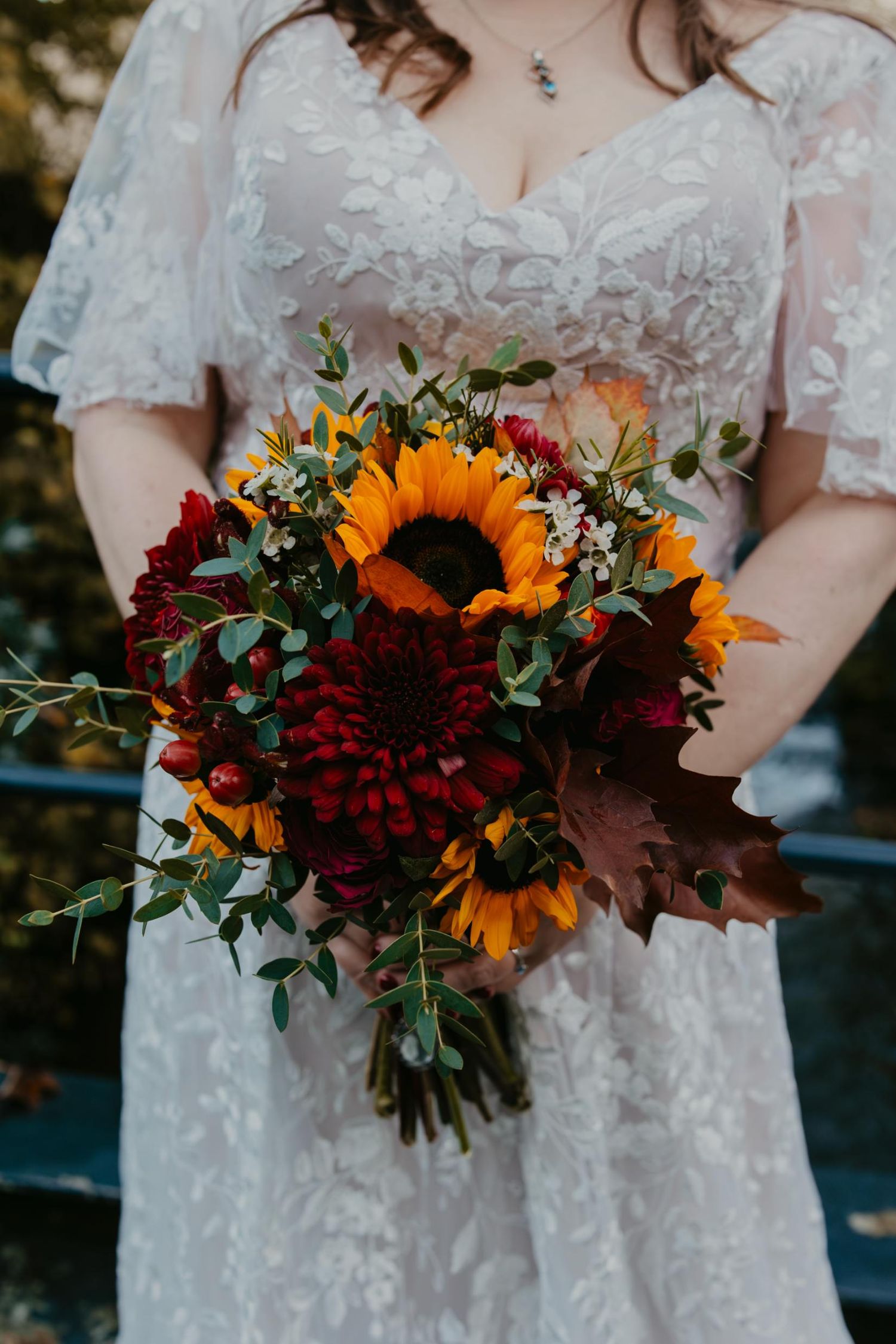 Bride in lace wedding dress holding autumn bouquet with sunflowers, red dahlias, and eucalyptus greenery.