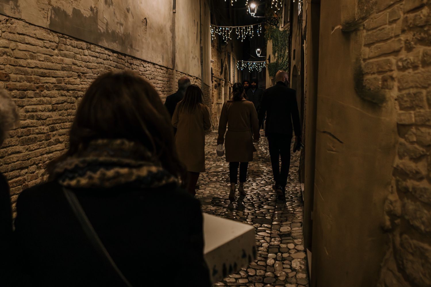 People walking through a narrow cobblestone alley at night, lit by string lights between old stone buildings.