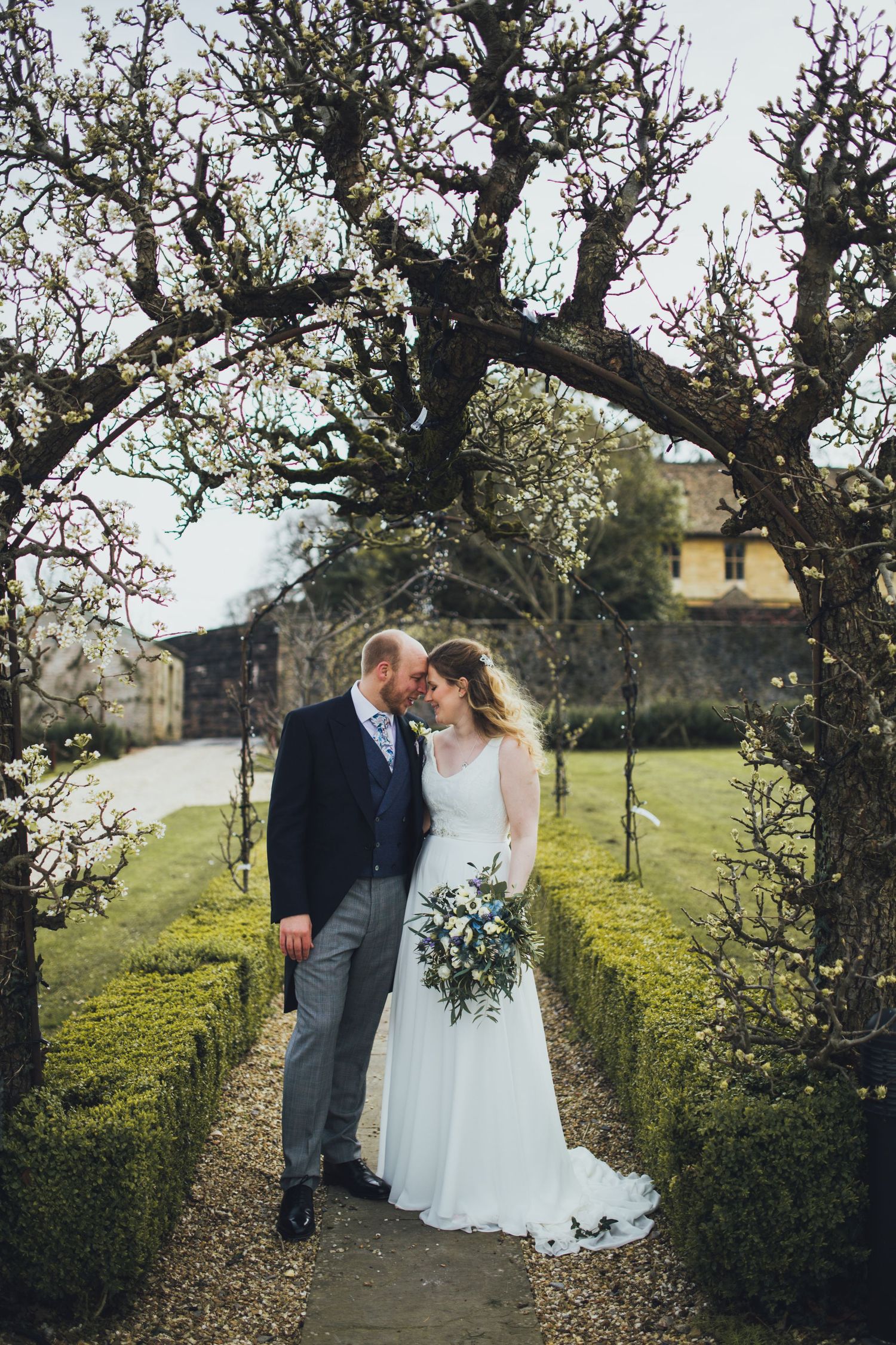 A couple shares an intimate moment on a garden path beneath flowering trees at a countryside venue.