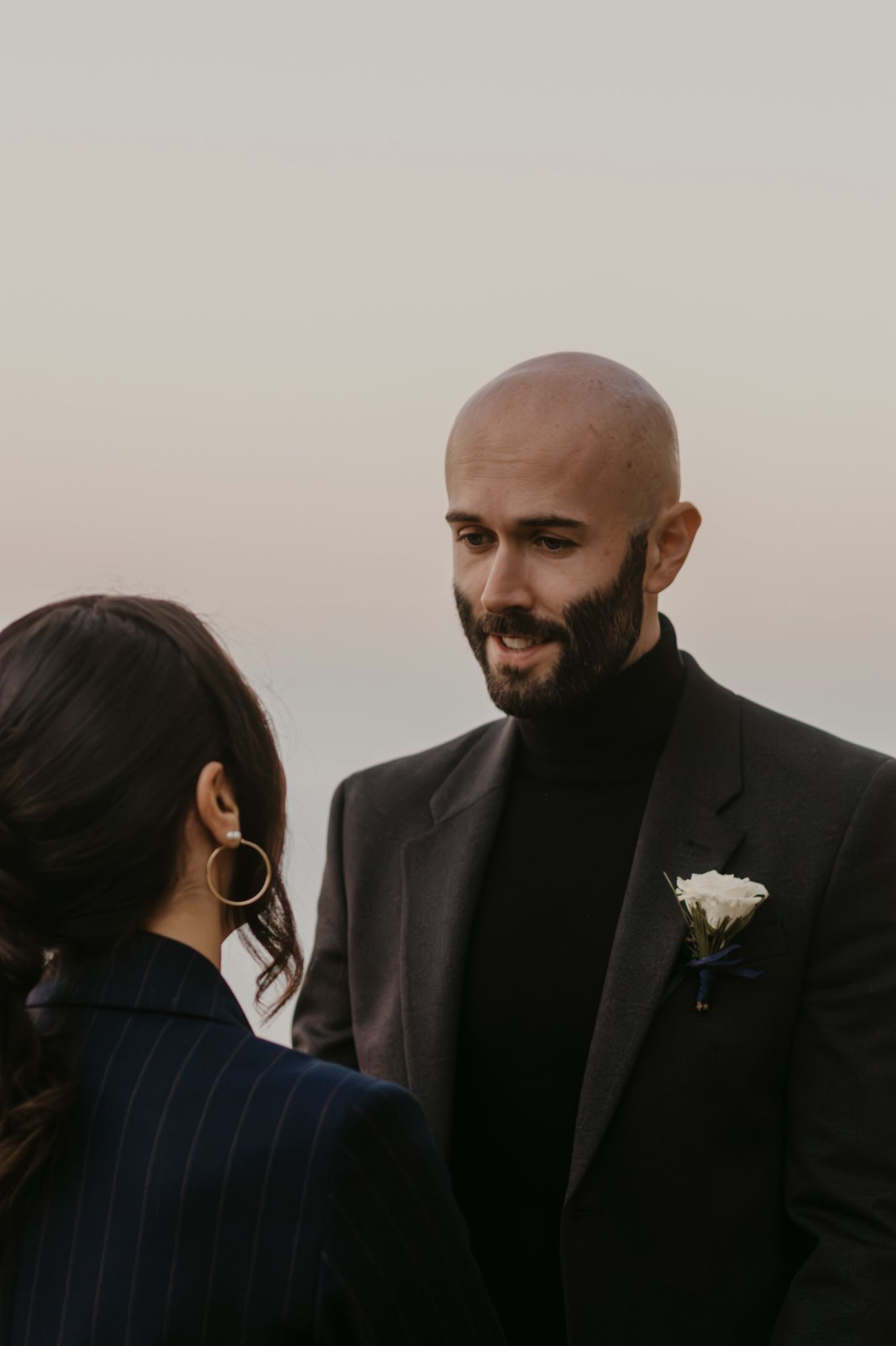 Groom with beard wearing dark suit and white boutonniere smiles at bride during outdoor wedding ceremony.
