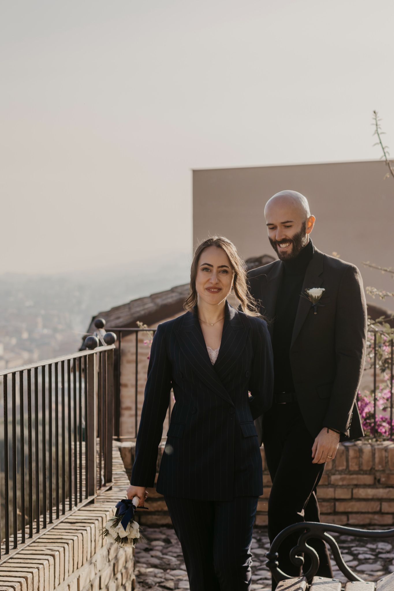 Couple in matching black suits walking on a terrace with a scenic cityscape backdrop on a sunny day.
