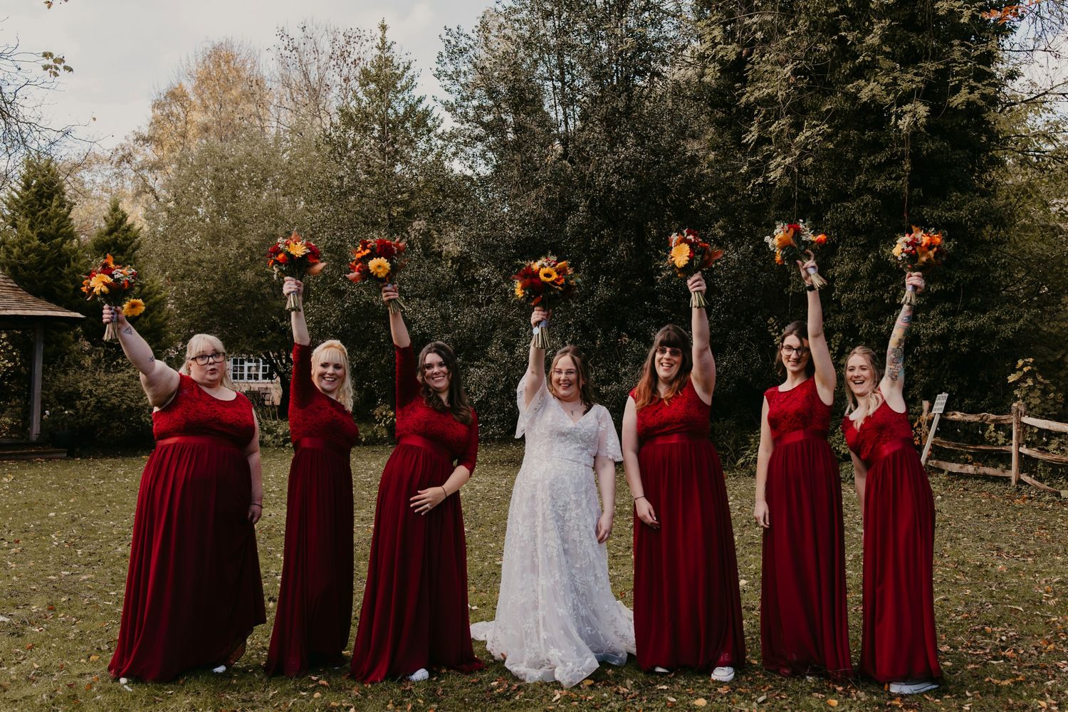 Bride in white lace gown with six bridesmaids in red dresses raising autumn bouquets outdoors at fall wedding.