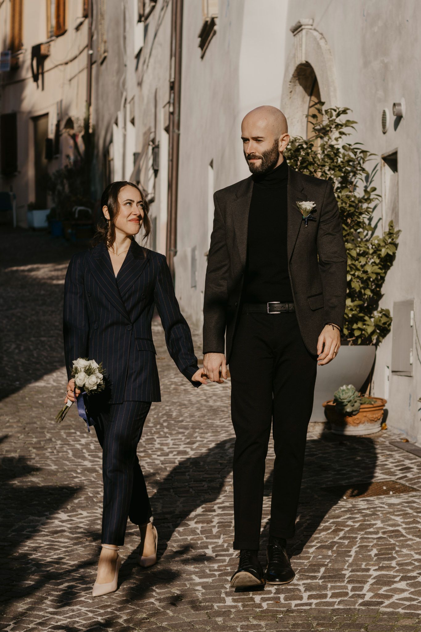 Couple in elegant dark suits holding hands and walking on cobblestone street, bride carrying white bouquet.