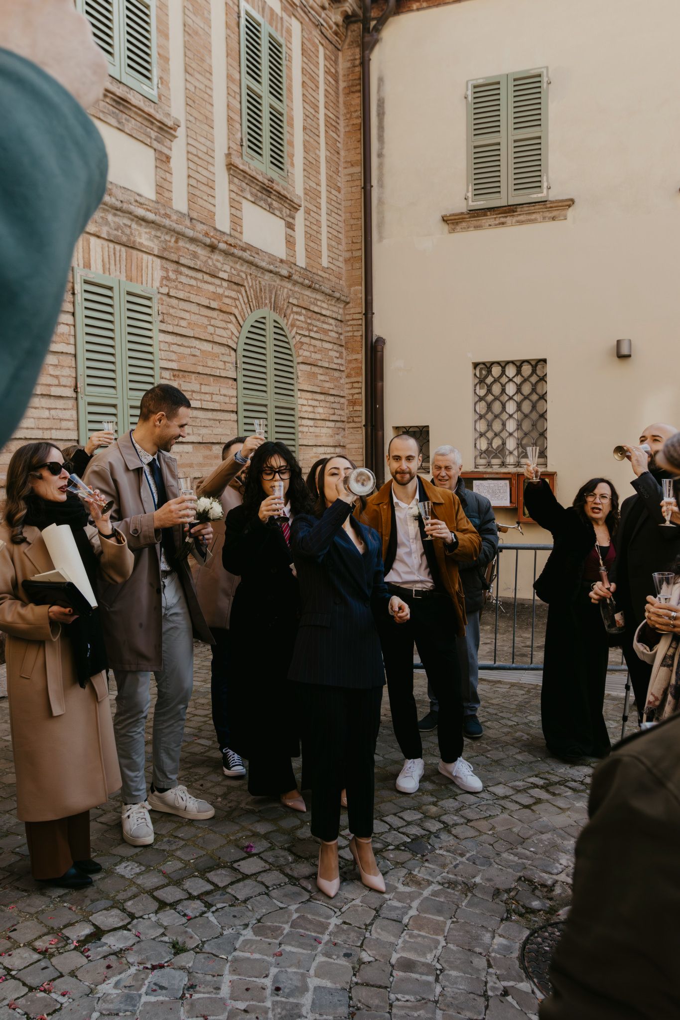 Group of elegantly dressed people celebrating outdoors in a cobblestone Italian street, raising glasses in a toast.