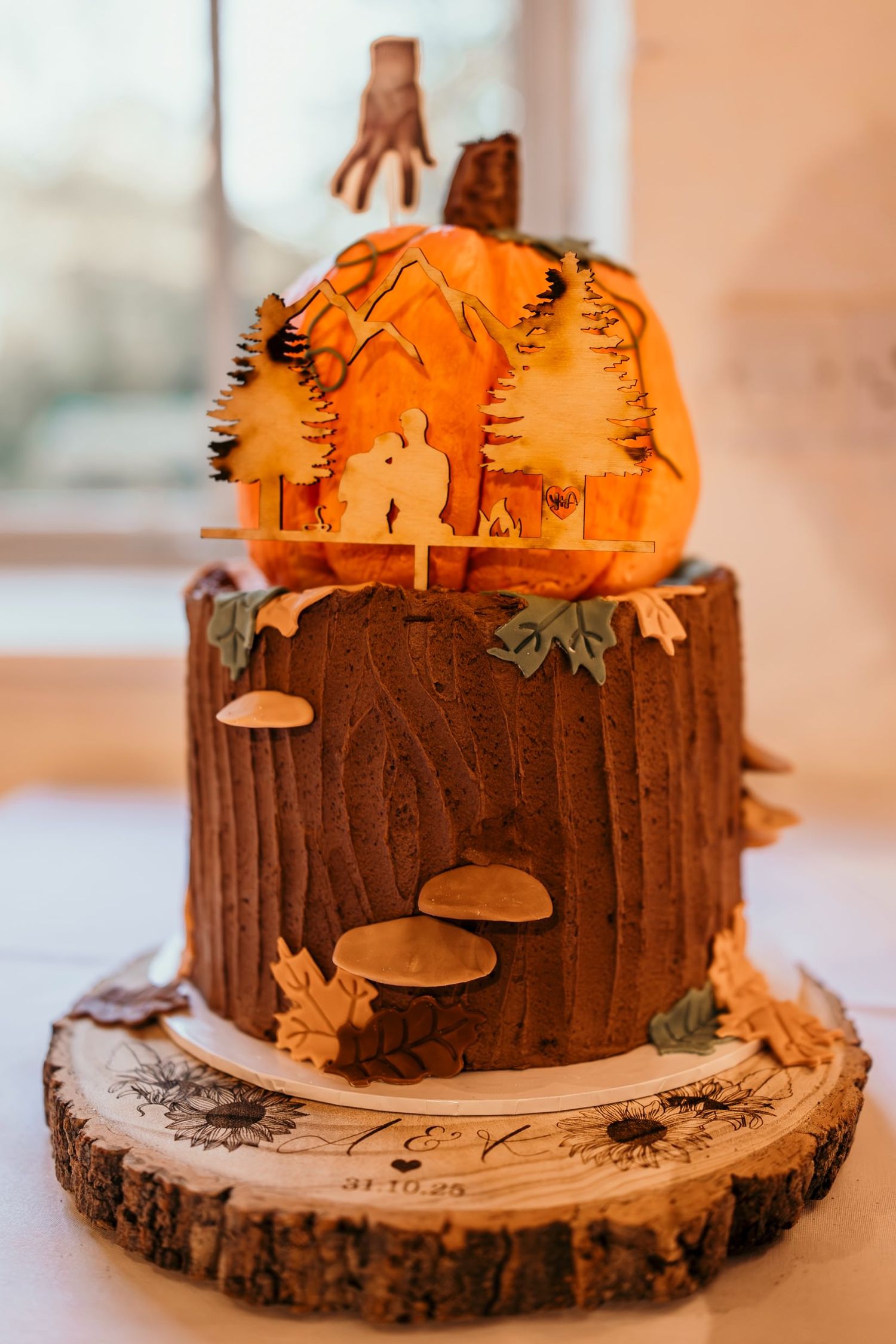 Two-tier Halloween wedding cake with pumpkin top, tree stump base, mushrooms, and silhouette topper on wood slice.
