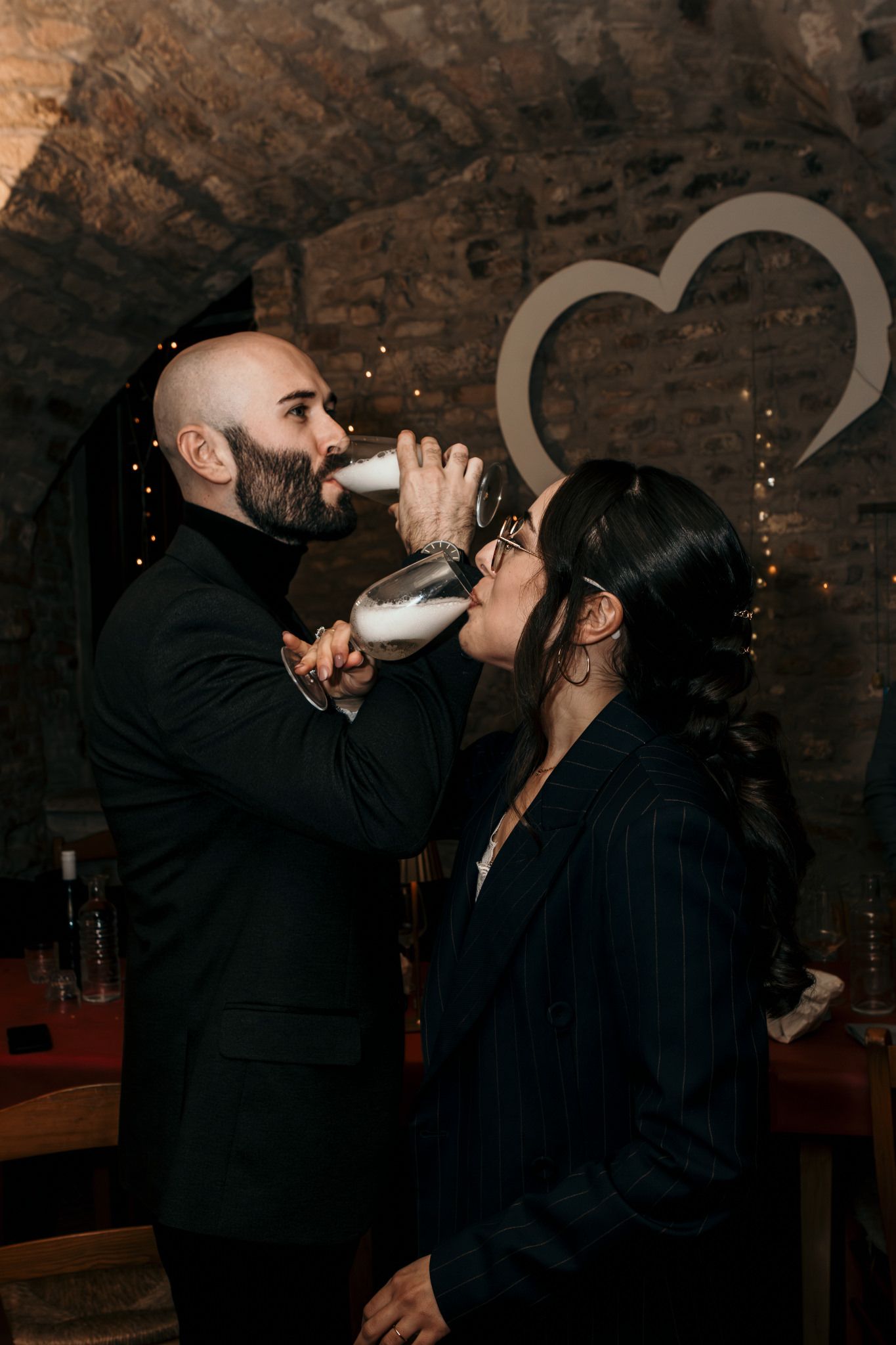 Couple sharing a drink from a large champagne glass in a romantic stone cellar venue with heart decor.