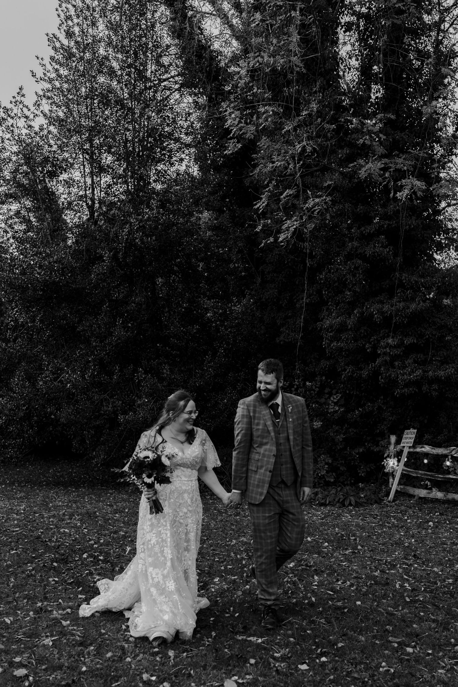 Black and white photo of a bride and groom holding hands, walking outdoors near lush trees on their wedding day.