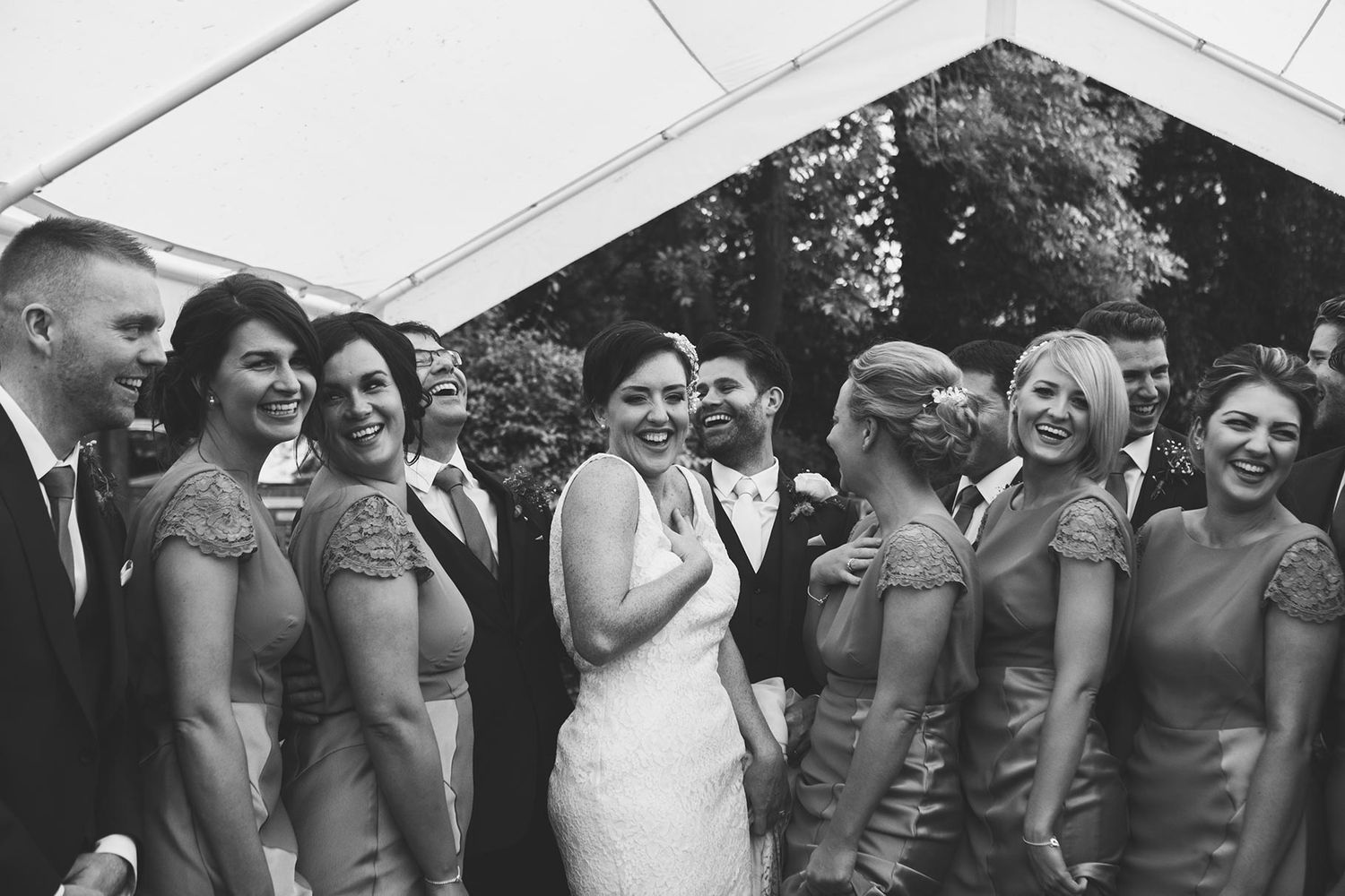 Black and white wedding photo of bride laughing with bridesmaids and groomsmen under a marquee tent.