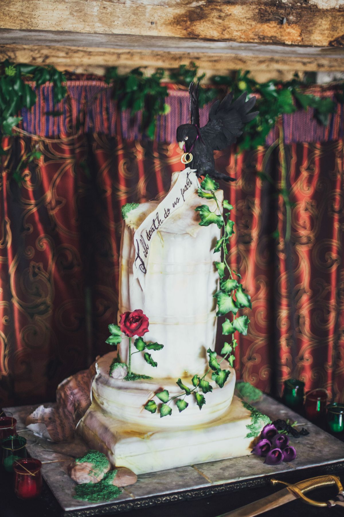 Elegant multi-tiered wedding cake with floral decorations, green vines, red roses, and black feather topper on ornate backdrop.