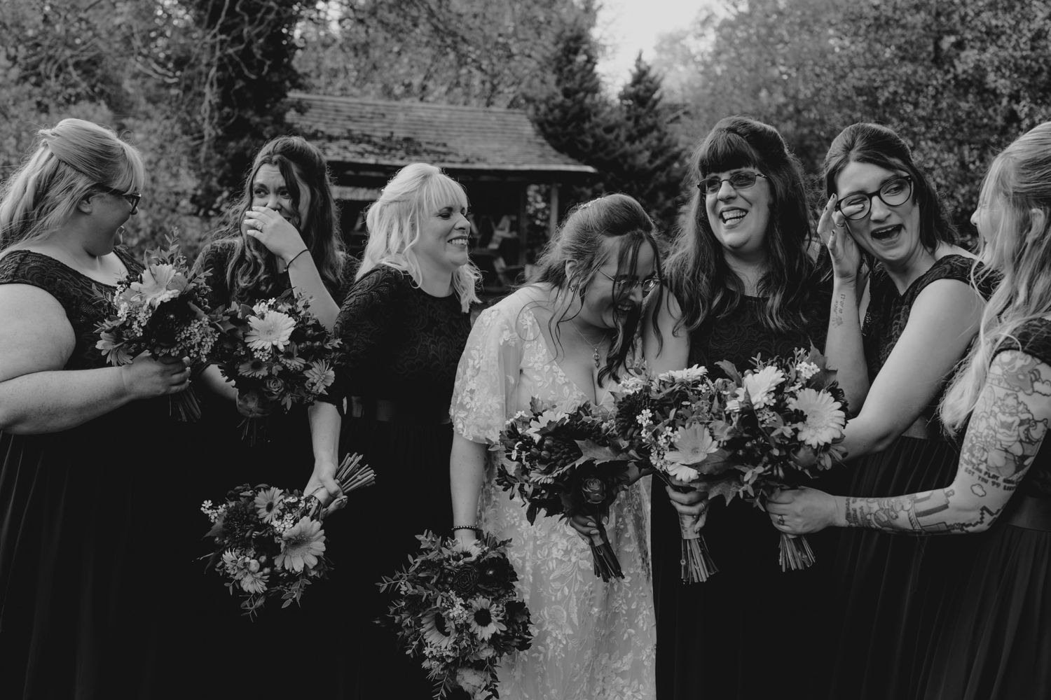 Black and white photo of a bride and bridesmaids in dark dresses laughing together while holding floral bouquets outdoors.