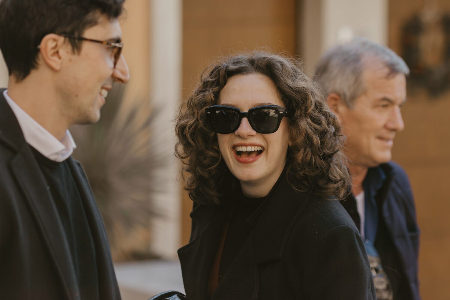 Three people smiling and laughing outdoors, woman in sunglasses and black coat enjoying a conversation.