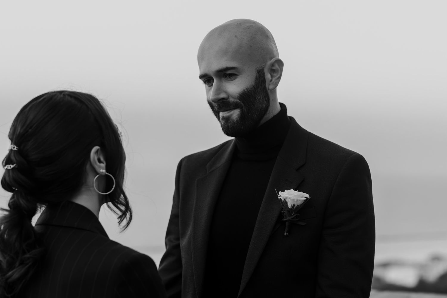 Black and white photo of a groom with beard in black tuxedo smiling at bride during outdoor wedding ceremony.