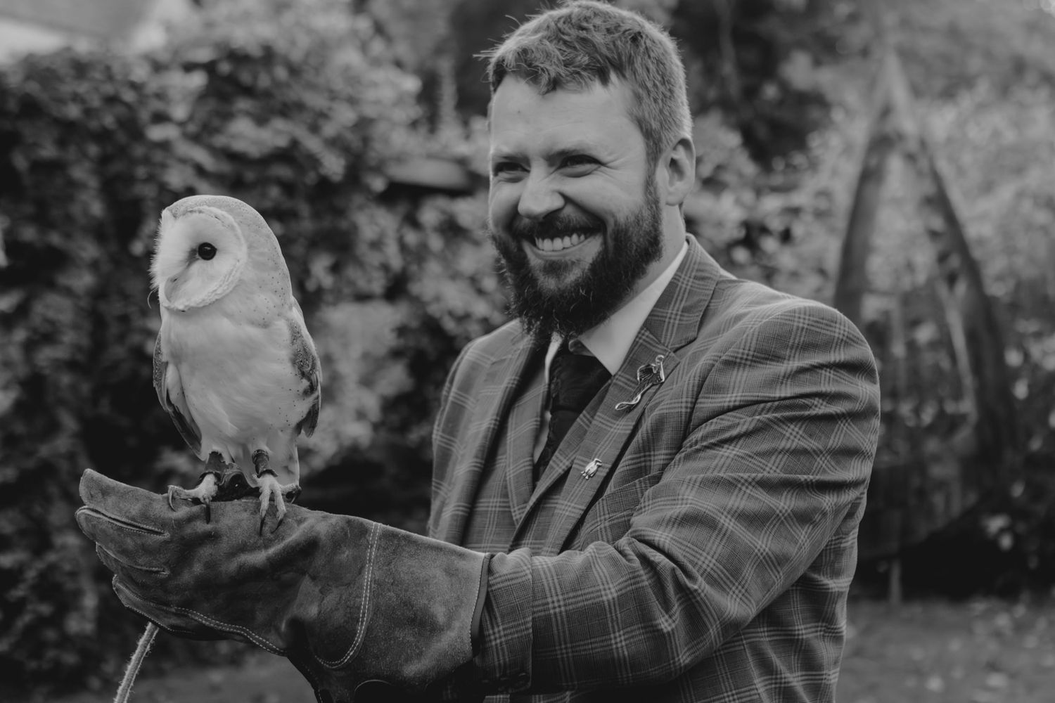 Smiling groom in tweed suit holding a barn owl on his gloved hand outdoors in black and white photo.