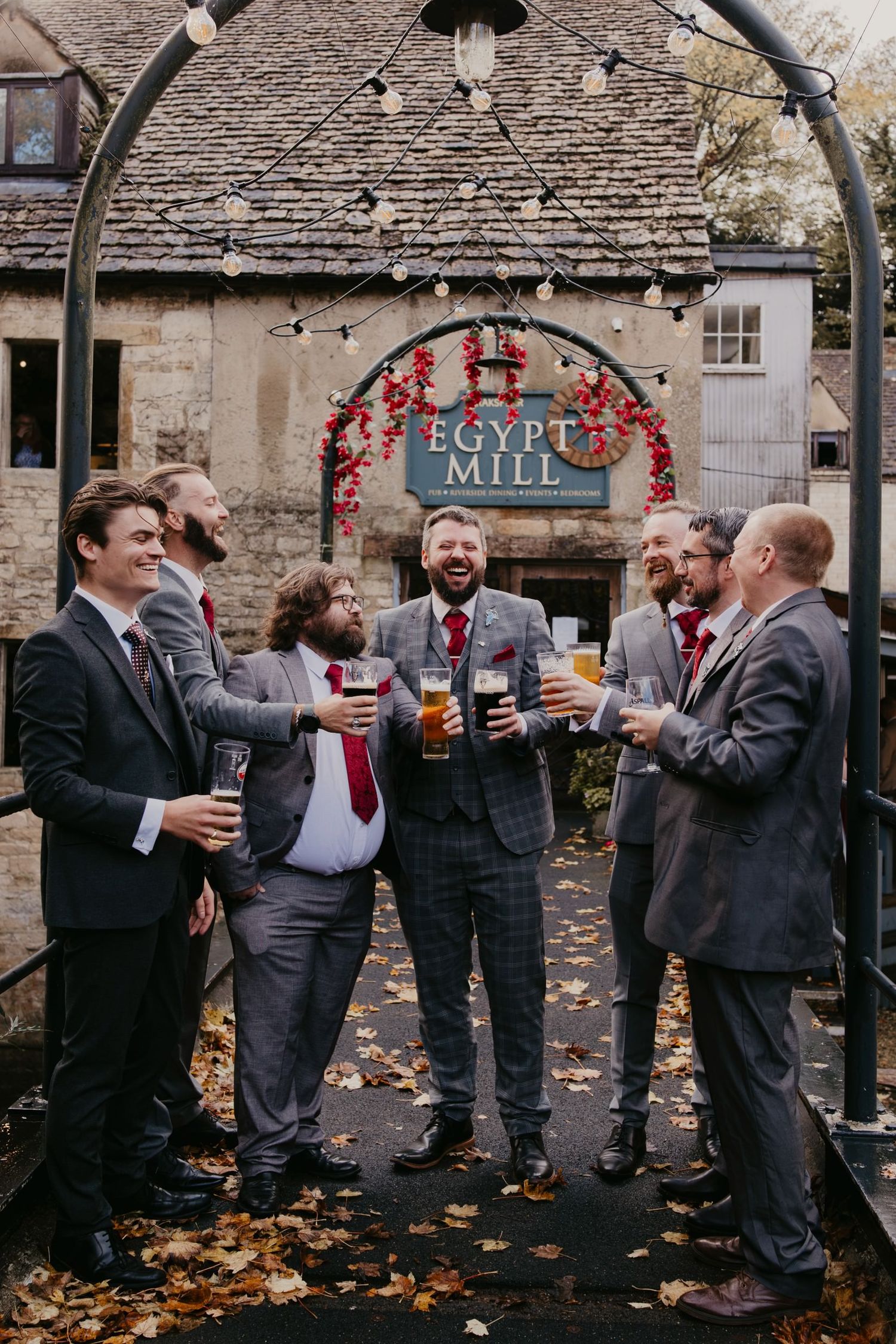 Groomsmen toasting with drinks outside Egypt Mill venue, wearing dark suits with red ties amid autumn leaves.