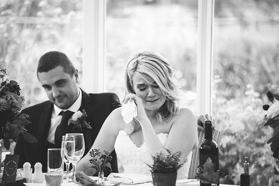 Emotional bride wiping tears at wedding reception while groom sits beside her at decorated table.