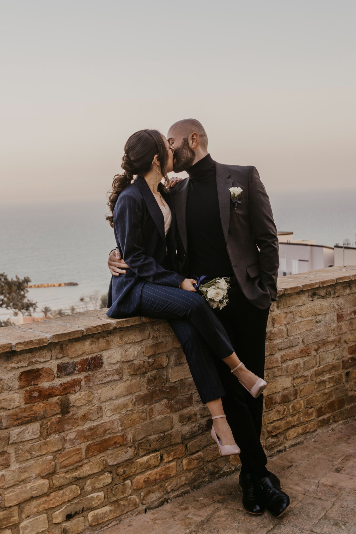Couple in elegant suits kissing on a stone wall with ocean view at sunset, holding a white bouquet.