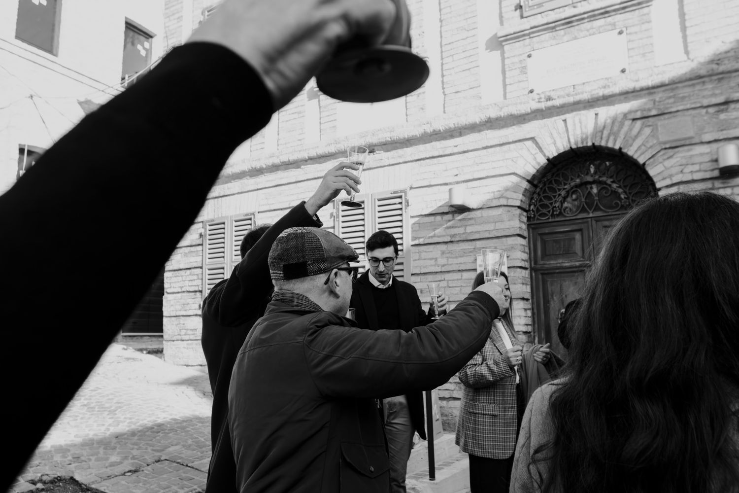 Black and white photo of a group of people raising glasses in a toast outdoors near a historic building.