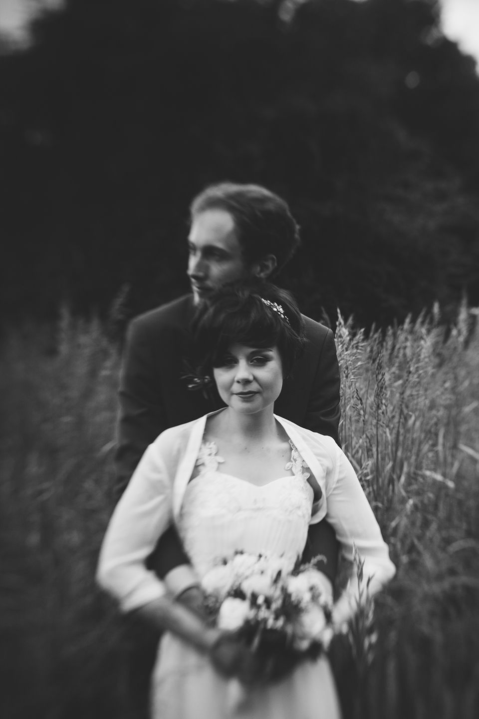 Black and white photo of a bride holding a bouquet standing before a groom in a field of tall grass.